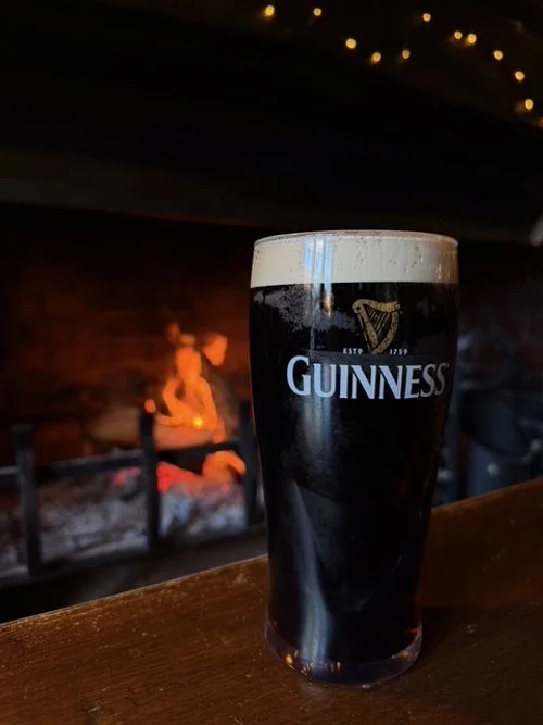 A pint glass of Guinness stout beer on a wooden bar counter with a fireplace in the background.
