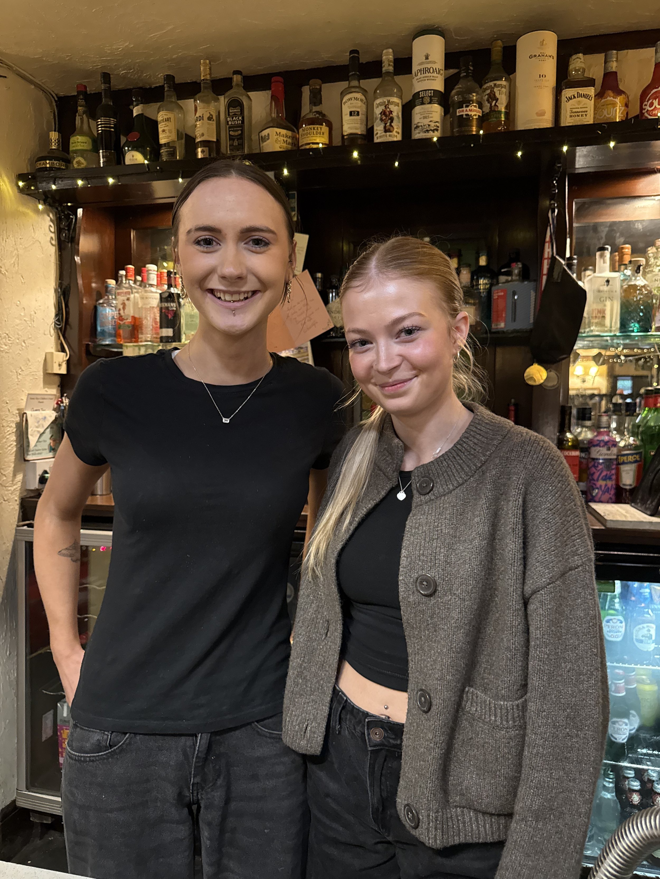 Two young women smiling in a bar with a bottles behind them on shelves.
