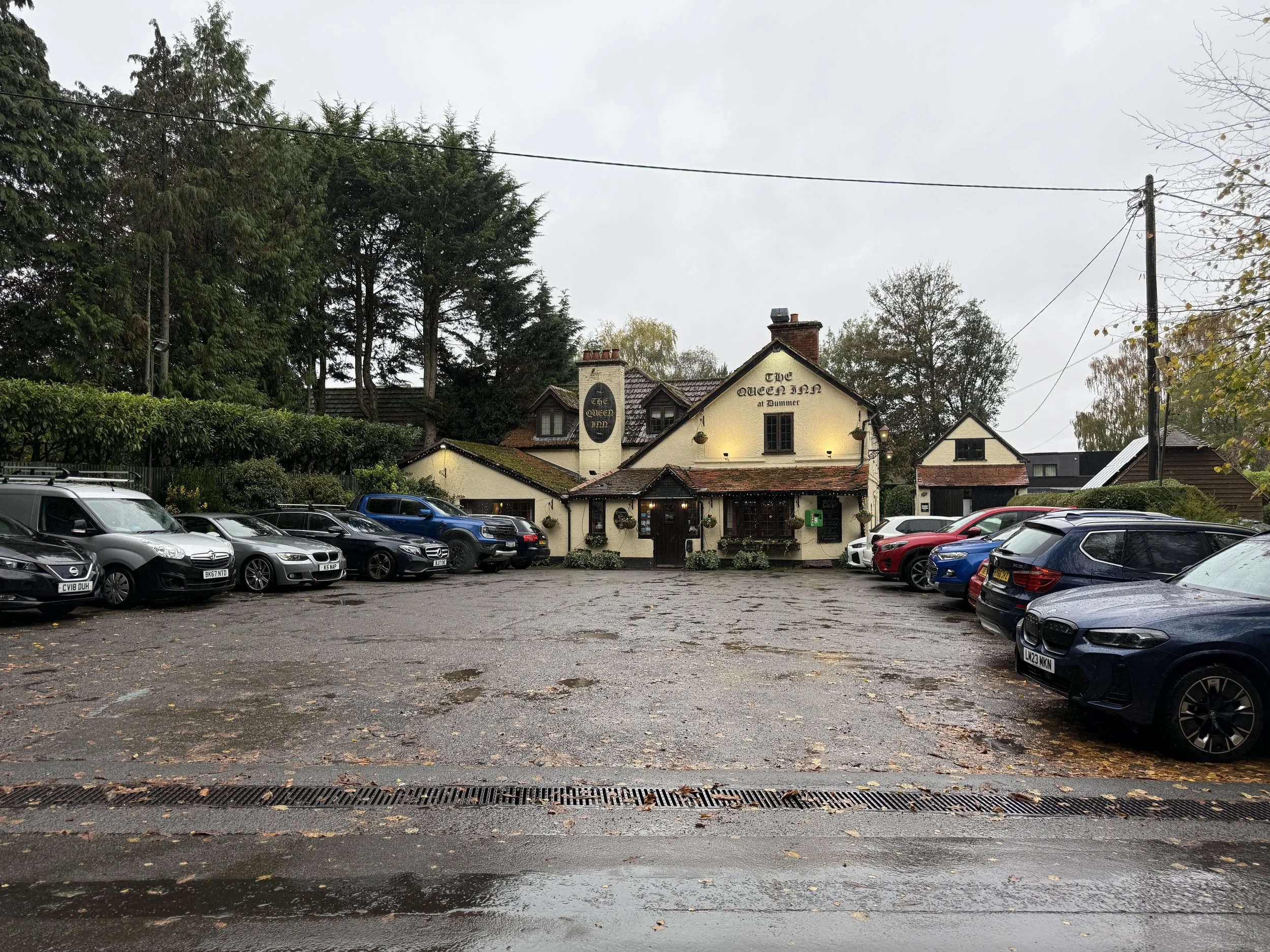 A parking lot in front of a pub called 'The Queen Inn at Tummer,' with cars parked on both sides, shortly after rain with wet pavement and fallen leaves, and a building with a tile roof and surrounding trees in the background.