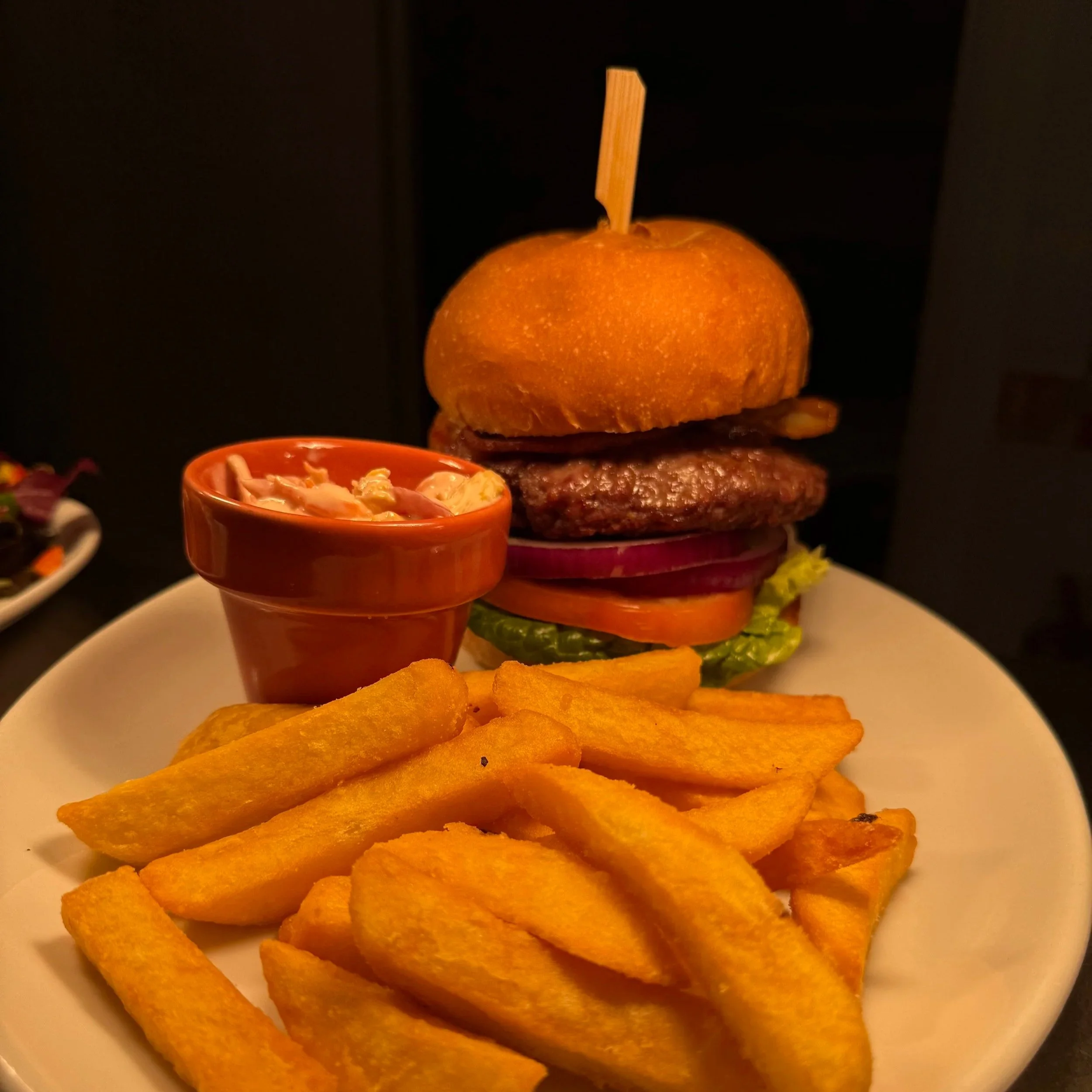 A gourmet burger with lettuce, tomato, onion, and pickles on a bun, served with a side of French fries and a small cup of coleslaw.