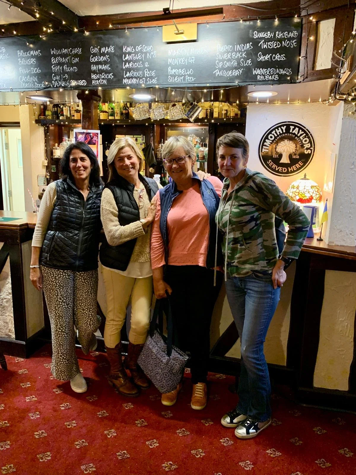 Four women standing together in a pub, smiling for the photo, with a bar in the background and a chalkboard menu above them.