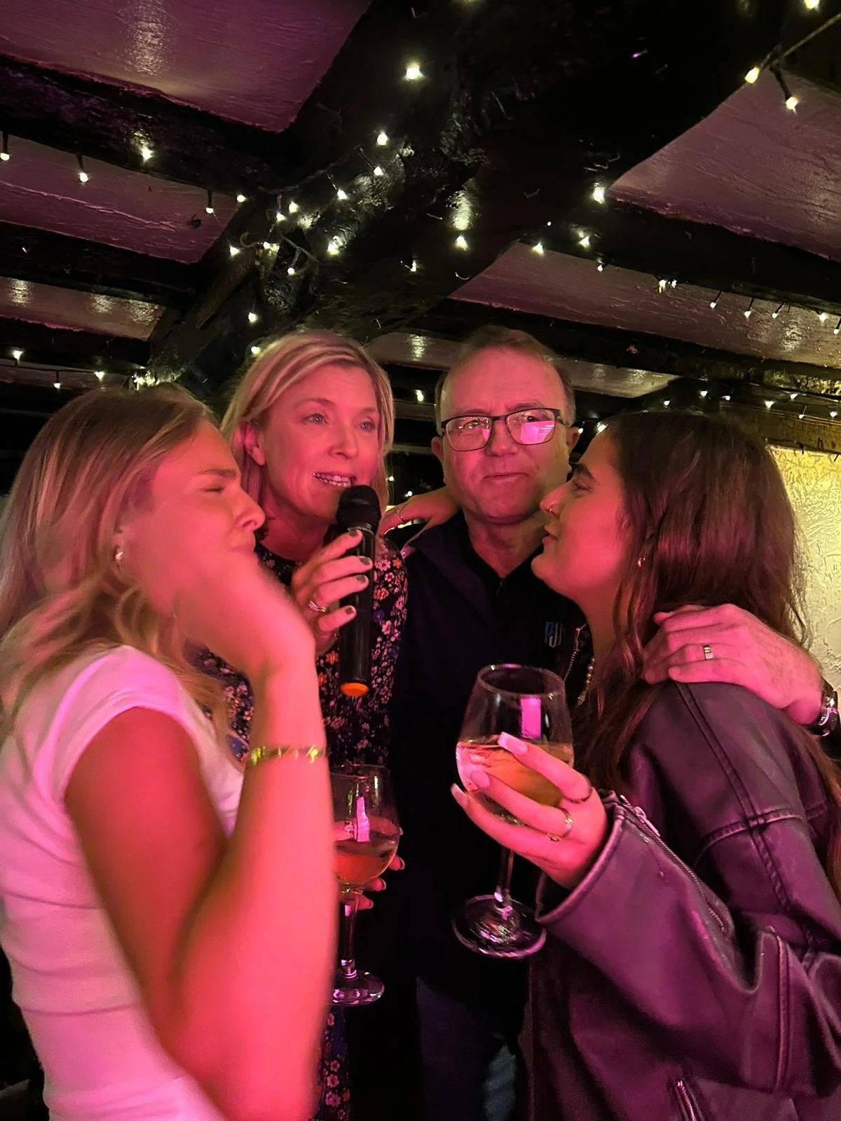 Group of four people at a party, two women holding wine glasses, one woman speaking into a microphone, a man in the middle, dimly lit with string lights on the ceiling.