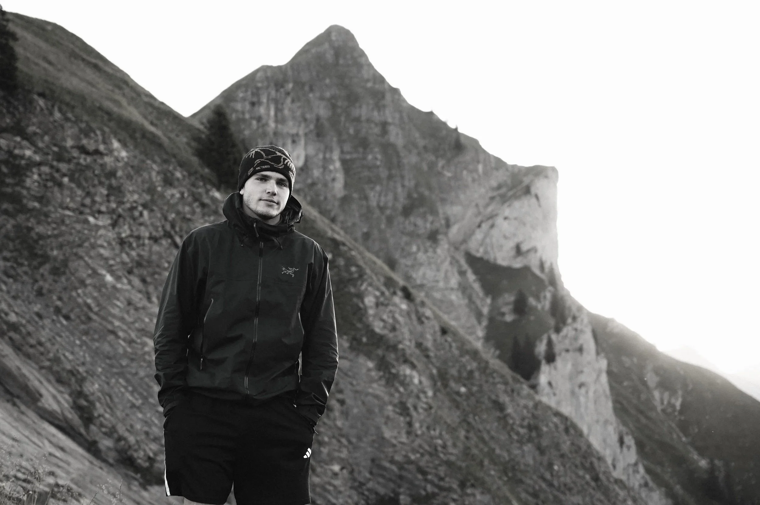 Junger Mann in schwarzer Arc 'teryx Jacke und Mütze steht vor bergiger Landschaft mit steil abfallenden Klippen, vermutlich im Gebirge.