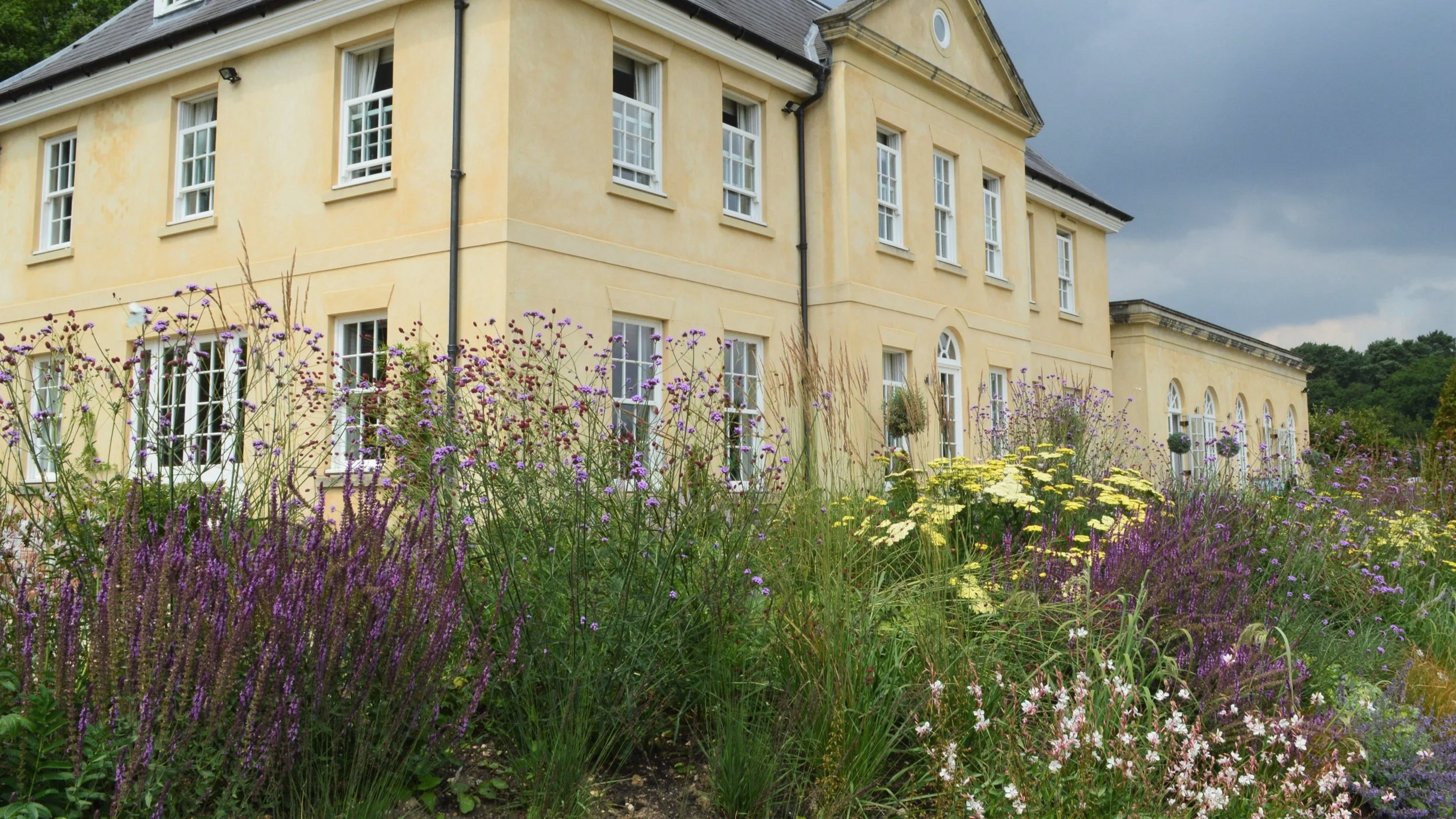 A yellow building with multiple windows and a garden filled with purple, yellow, and white flowers in the foreground, under a cloudy sky.