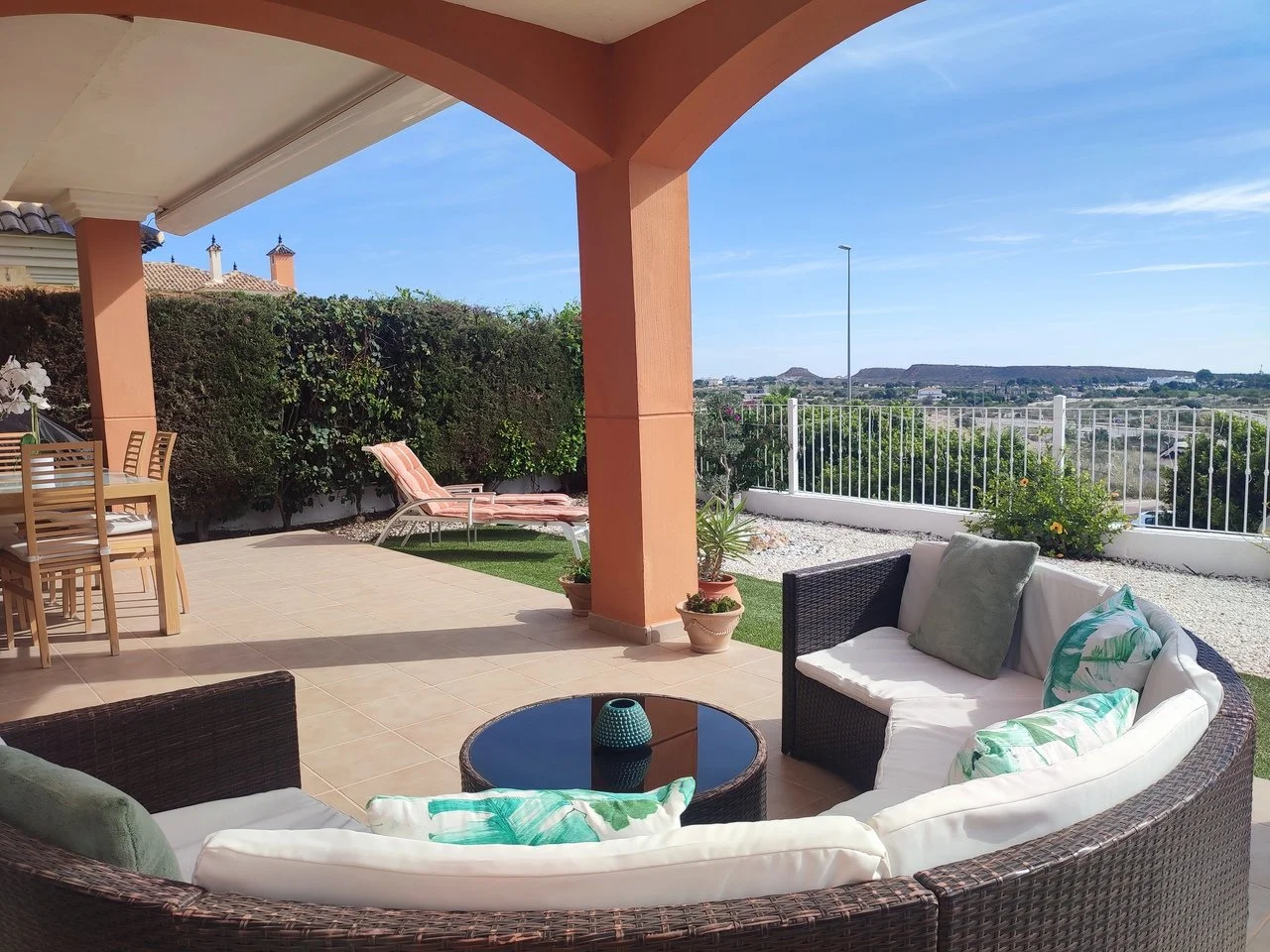 Outdoor patio with wicker sectional sofa, cushions, small black table, patio table with chairs, sun loungers, potted plants, and scenic view of hills and sky in the background.