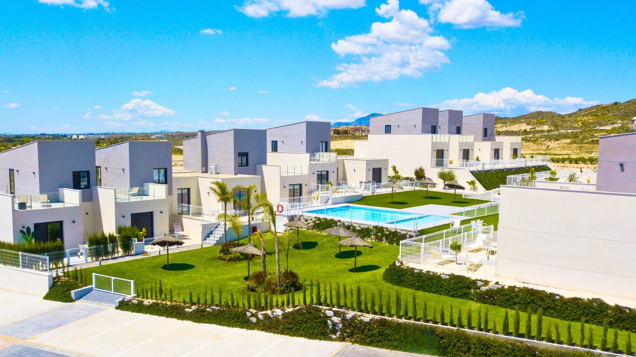 A modern residential complex with white buildings, a swimming pool, and a well-maintained green lawn with small trees and umbrellas, set against a backdrop of hills and a blue sky with clouds.