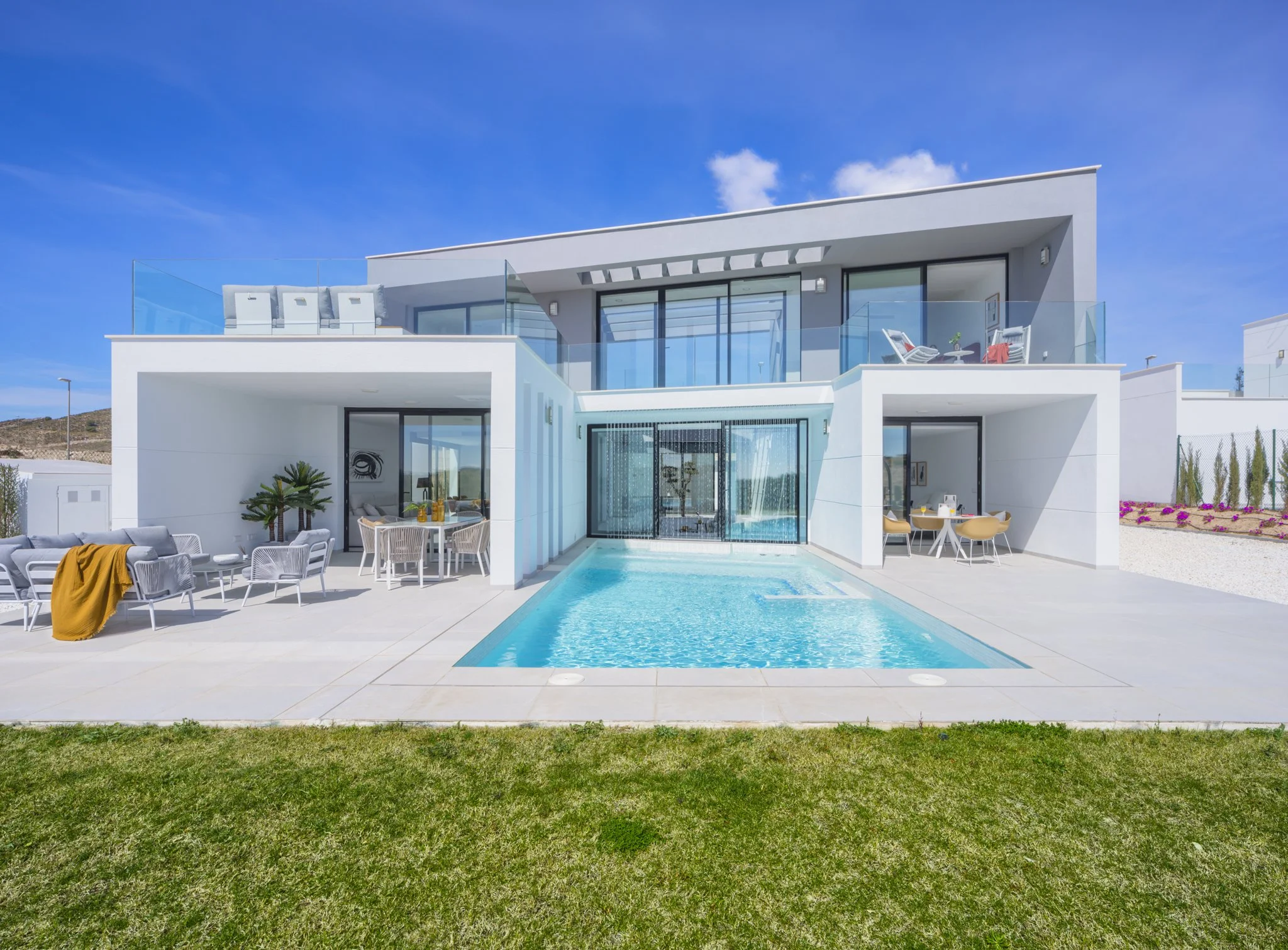 Modern white house with large glass windows and a swimming pool in front, featuring outdoor seating areas with chairs and tables under a bright blue sky.