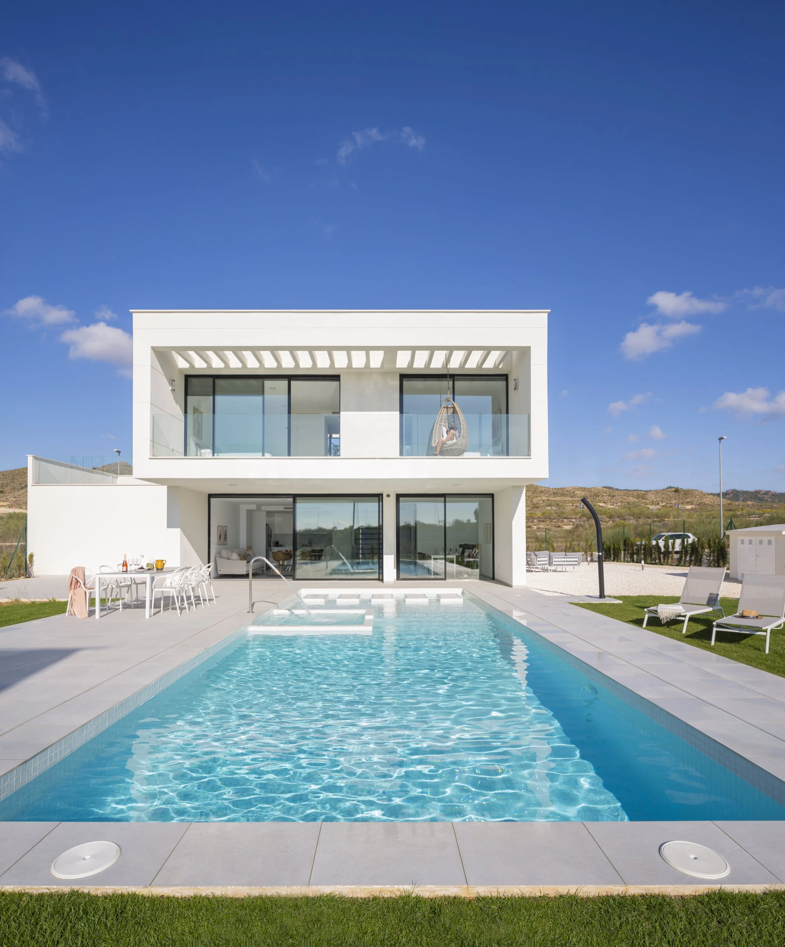 Modern white house with a swimming pool in the foreground, outdoor seating area on the left, lounge chairs on the right, and a person hanging in a hammock on the second-story balcony, under a clear blue sky.