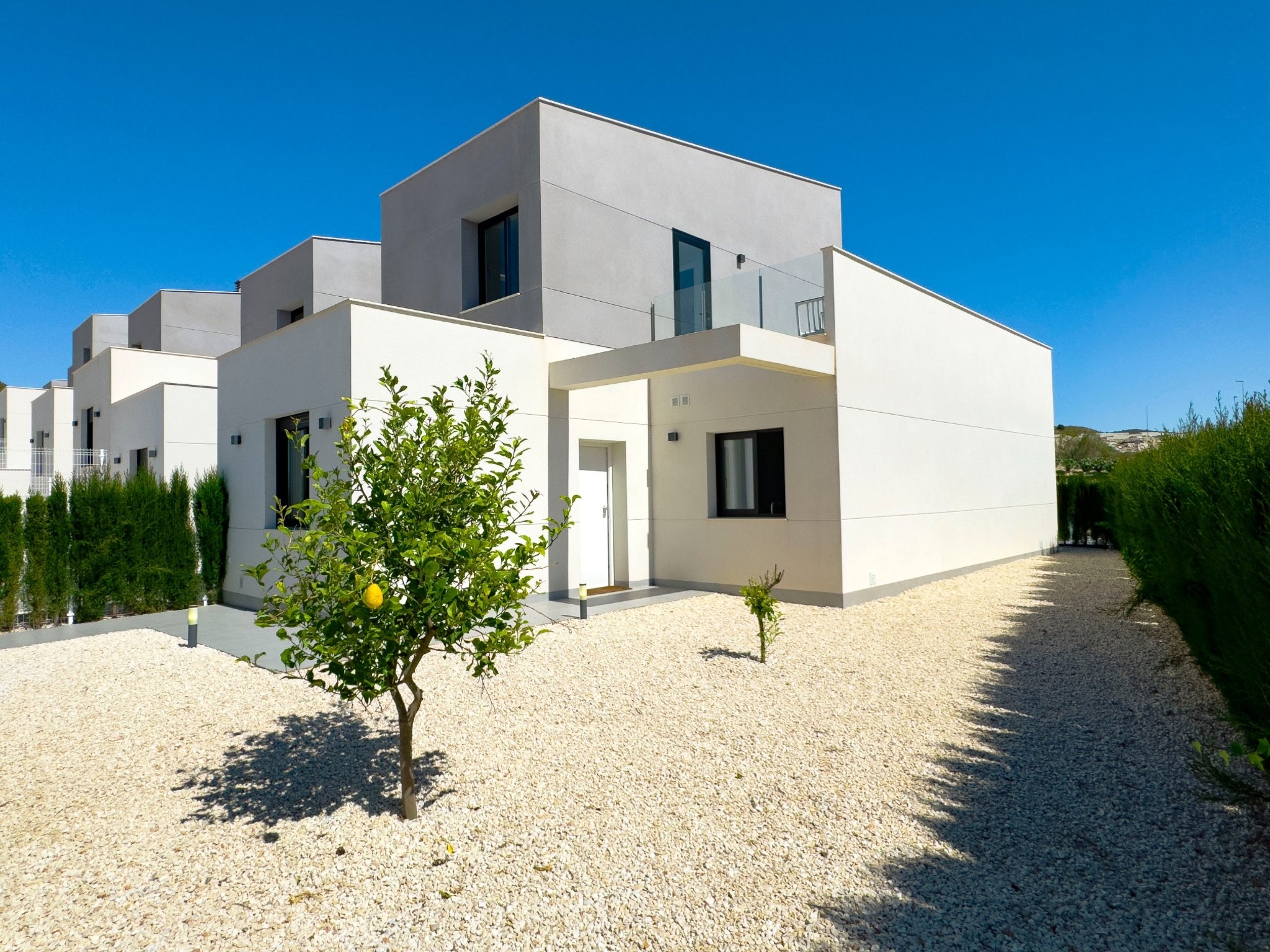 Modern white multi-story residential building with minimalist design, gravel front yard with small lemon tree, green hedges, and clear blue sky.