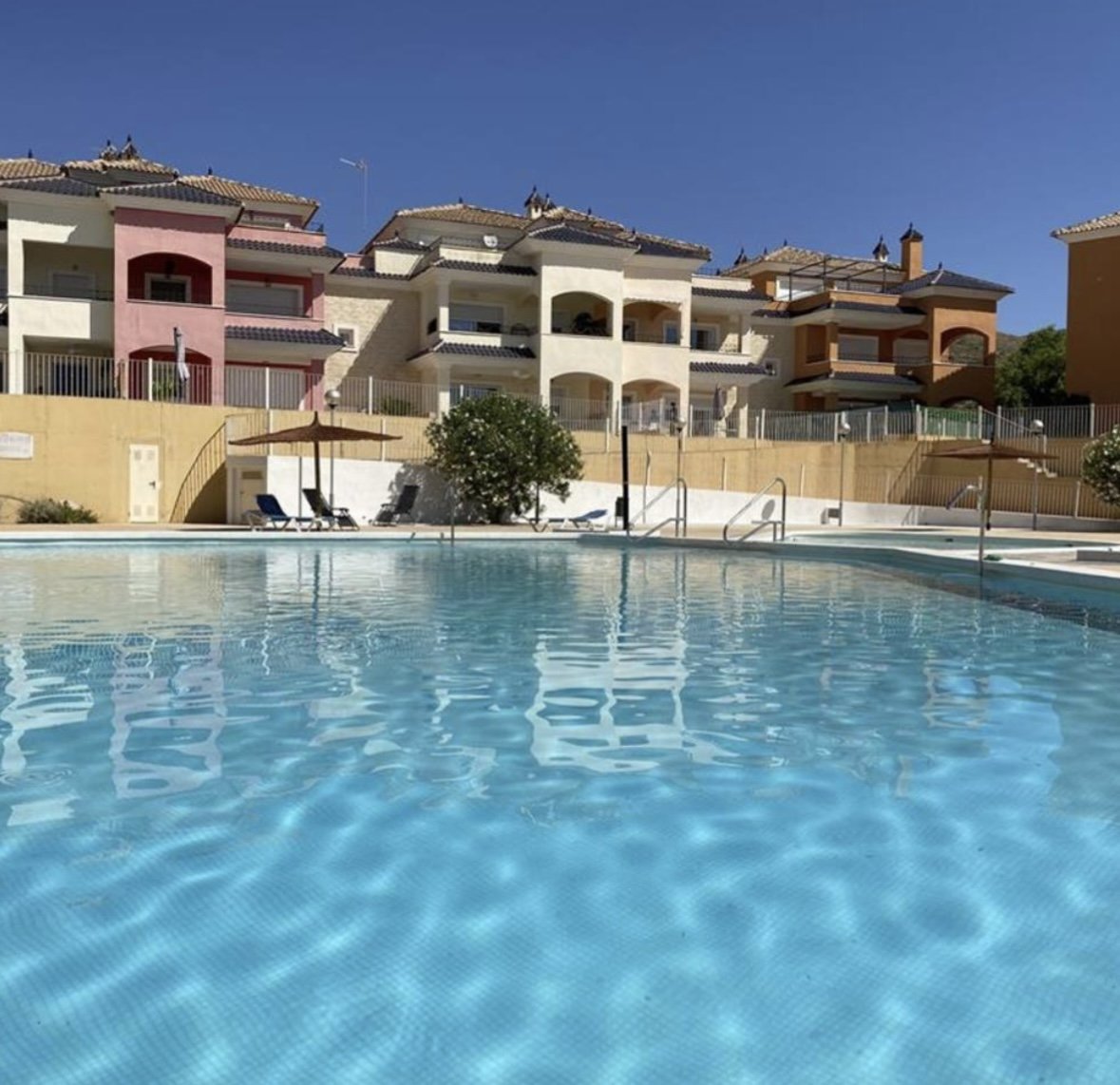 A swimming pool with clear blue water, patio chairs, and an umbrella, surrounded by colorful residential buildings under a clear blue sky.