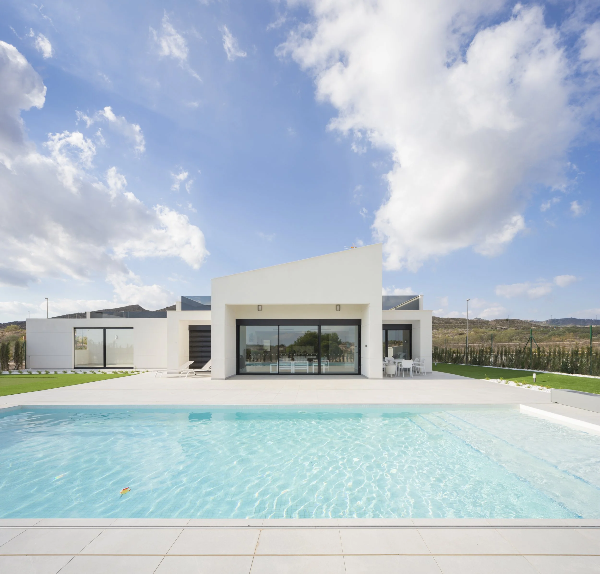 Modern white house with large glass sliding doors, a swimming pool in the foreground, outdoor furniture, and a partly cloudy sky with mountains in the background.
