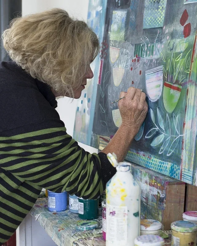 An older woman with curly blonde hair painting on a canvas with pots of colorful paints on a table in front of her.