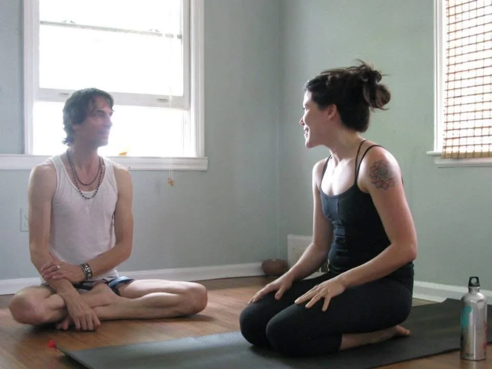 Marsha McNeight sitting on her mat next to David Garrigues, certified Ashtanga Yoga teacher. They are smiling at each other.