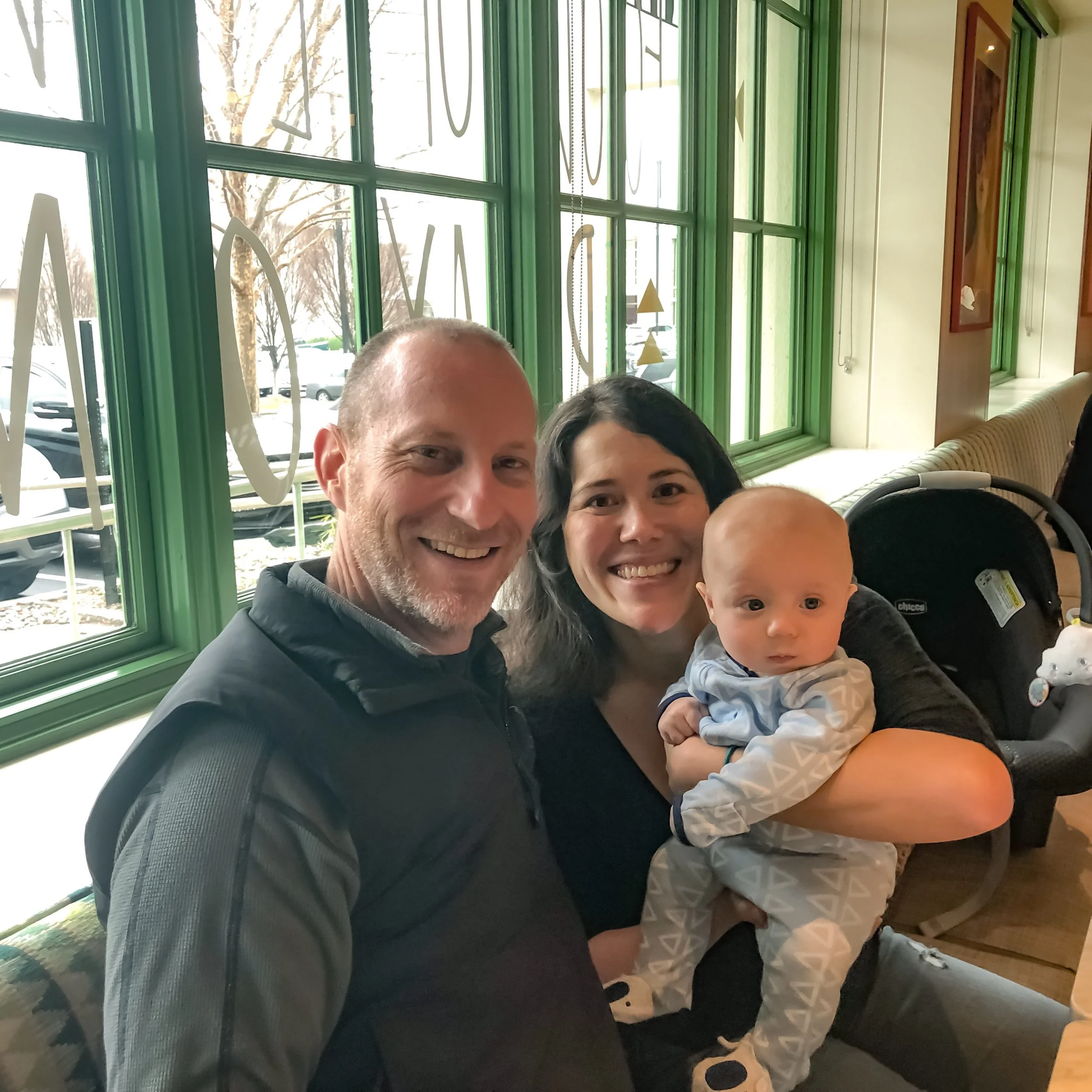 Marsha McNeight and her young son smiling with David Keil, Authorized Level 2 Ashtanga teacher, sitting inside a cafe with large green window frames and outside visible parking lot and trees.