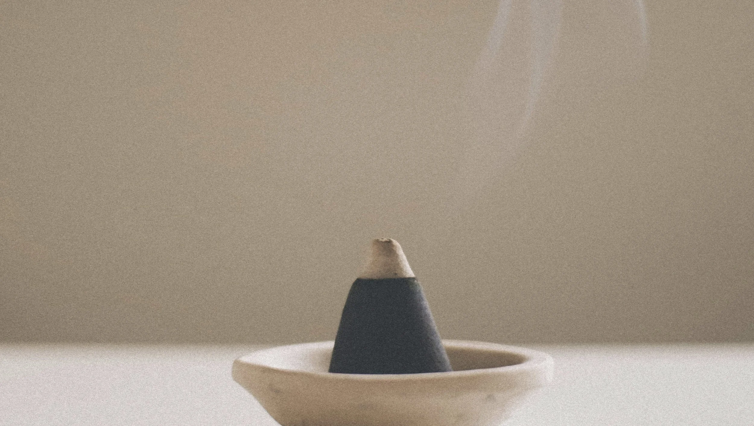 A charcoal grey incense cone burning with ash on the tip, placed in a white ceramic dish on a white surface with a plain beige background.