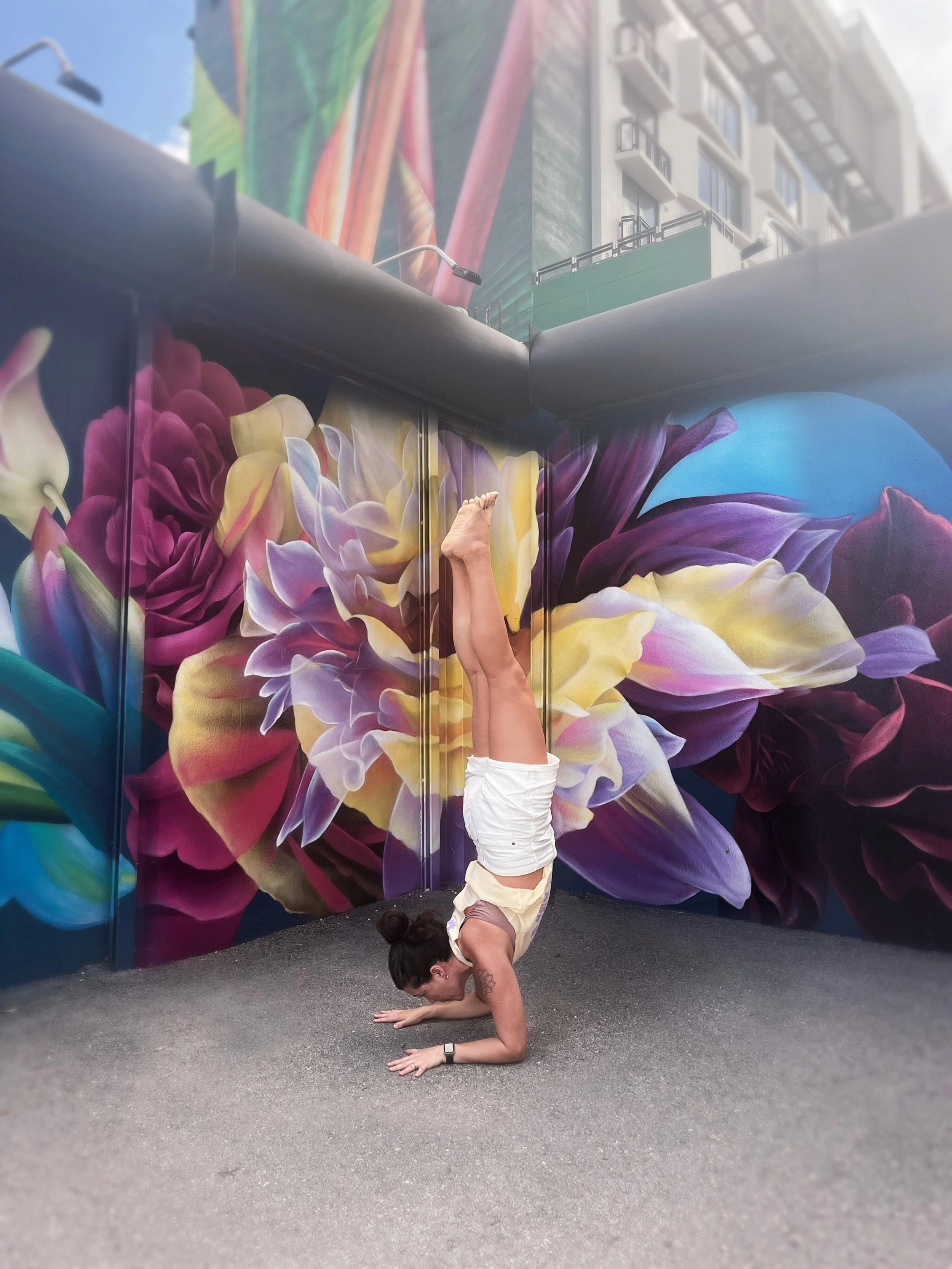 A woman performing a headstand yoga pose against a vibrant mural of large, colorful flowers and a blue globe on a city street.
