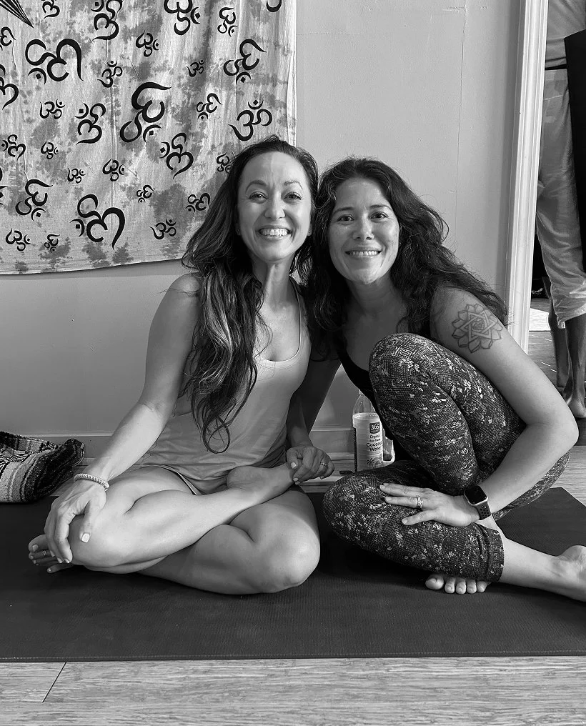 Two women sitting cross-legged on yoga mats and smiling. One woman has long wavy hair and is wearing a tank top, the other has curly hair, a tattoo on her arm, and is wearing patterned leggings. Behind them is a curtain with Asian characters and a person standing partially visible.