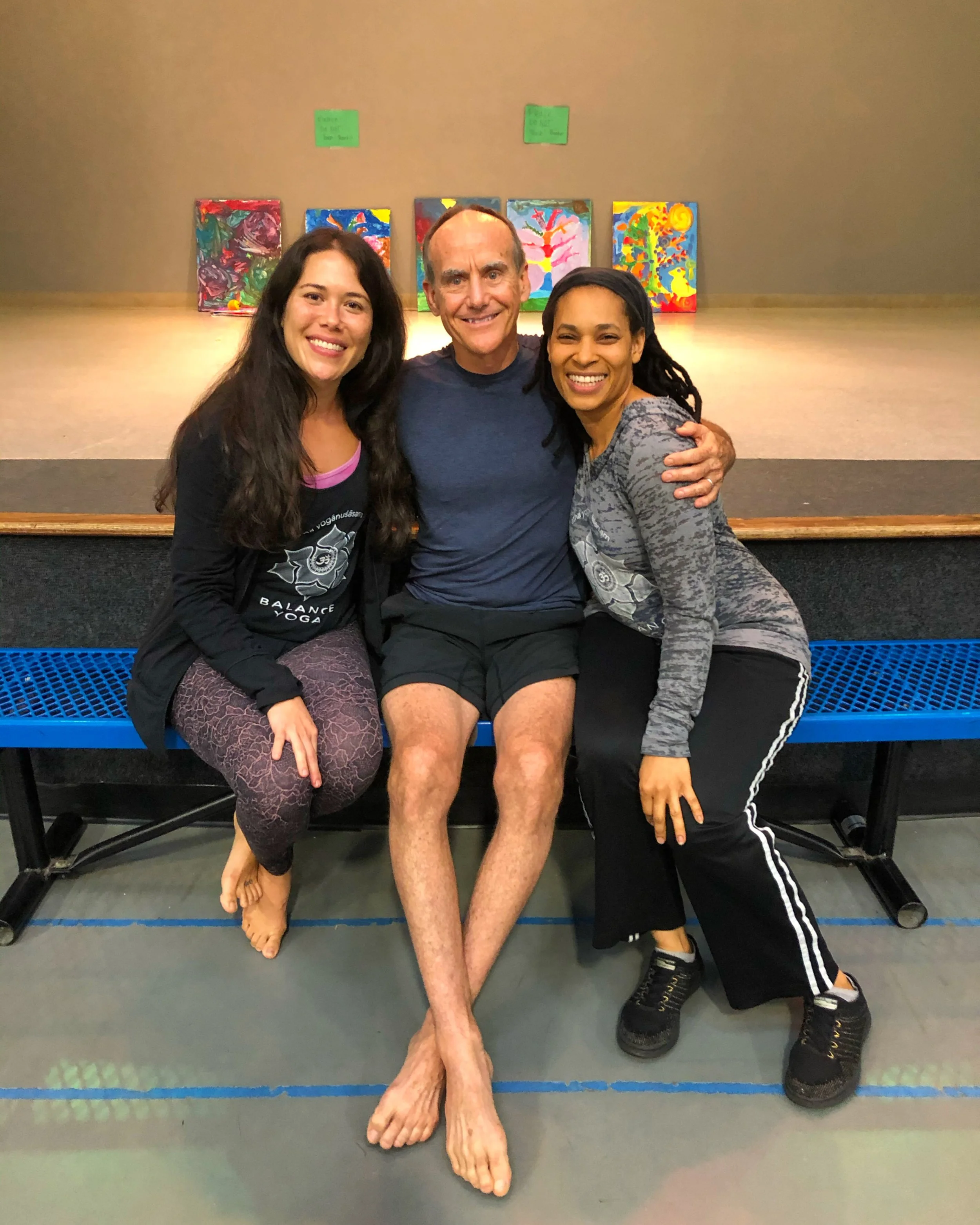 Marsha McNeight with David Swenson, Ashtanga Yoga teacher, and another student, sitting close together on a blue bench, smiling at the camera.  Behind them is a stage with colorful abstract paintings displayed on the wall.