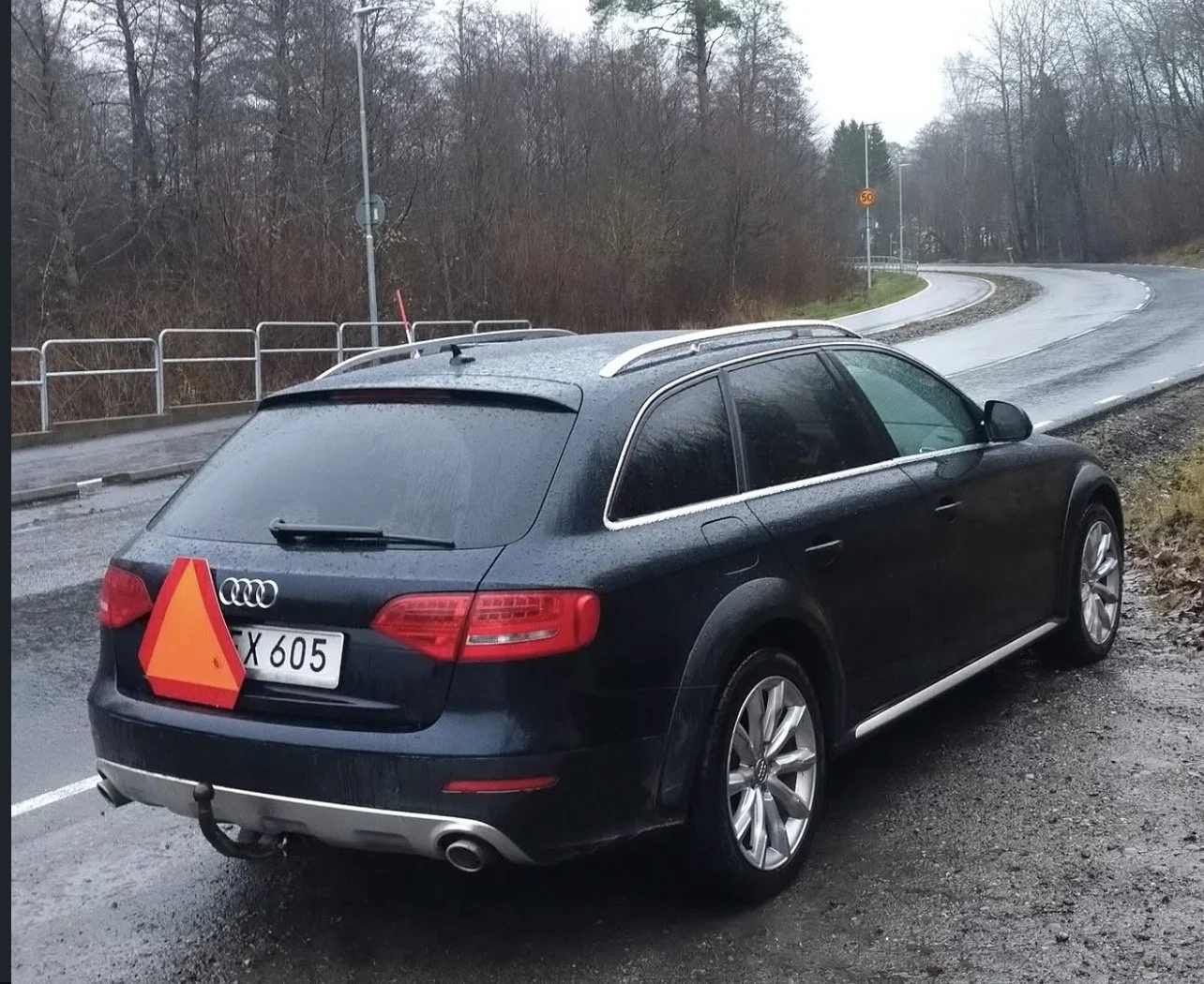En svart Audi station wagon A-traktor med ett orange triangel på bakluckan, parkerad på en väg med kurva, omgiven av träd och ett järnstaket, på en regnig dag. Nyregistrerad A-traktor. Nyombyggd a-traktor.