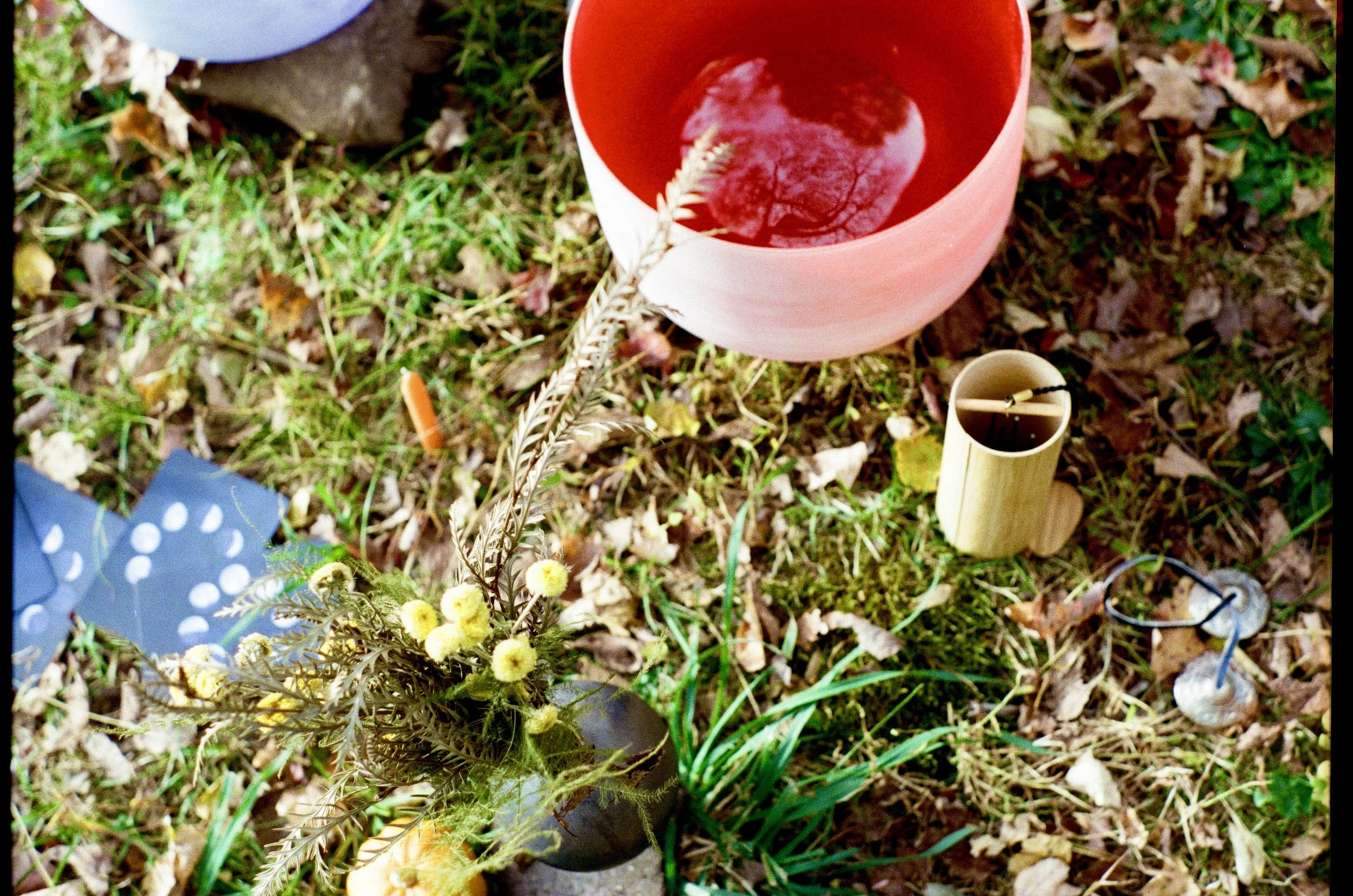 Memorial setup with a pink candle, a small bouquet of yellow and brown flowers, a bamboo candle holder, and small decorative items on a grassy and leaf-covered ground.