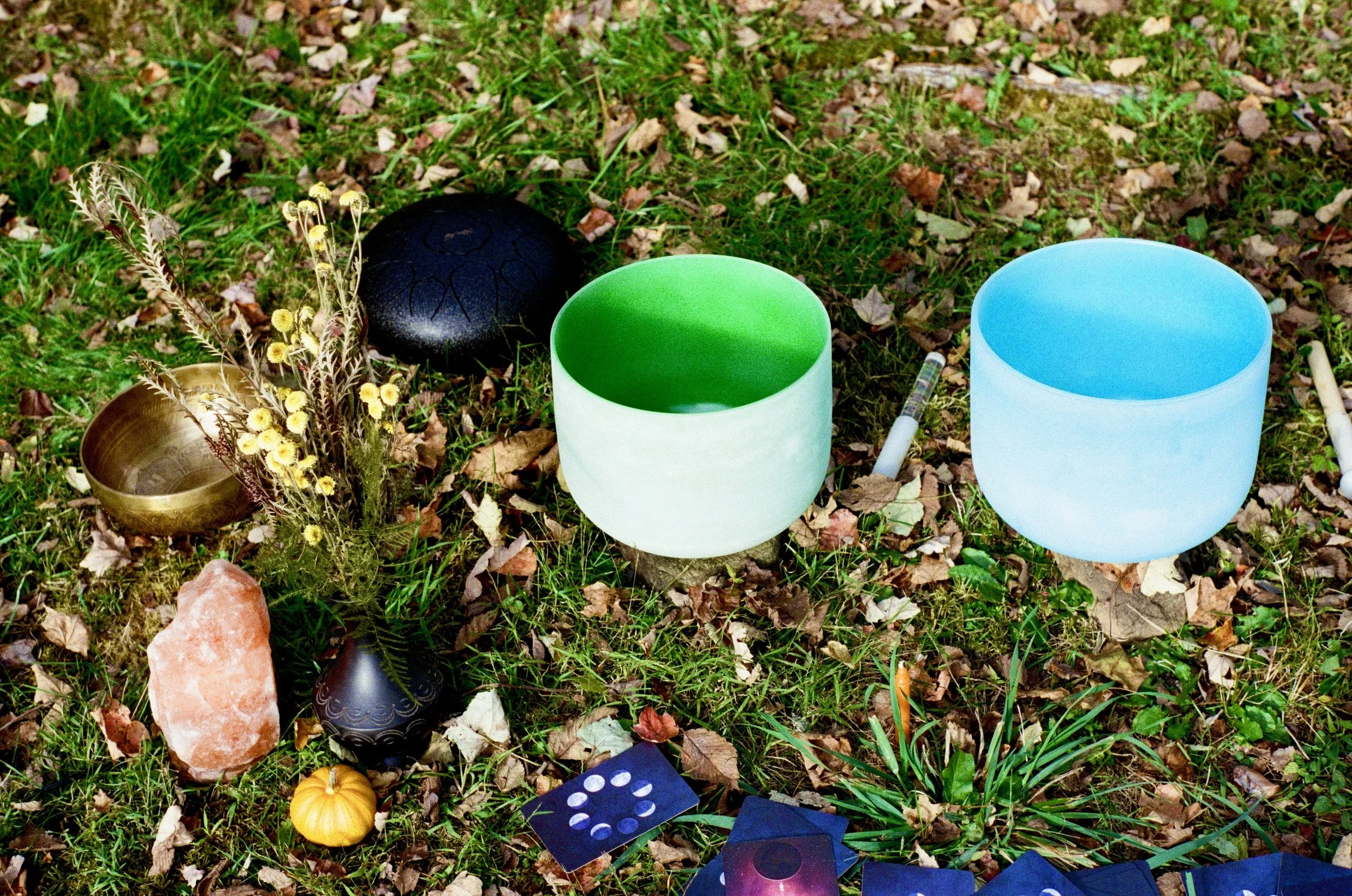 A collection of musical and spiritual items placed on grass, including two crystal singing bowls, a black gong, a bronze bowl, a small plant with yellow flowers, a pink salt crystal, a miniature pumpkin, tarot cards, and a white and a black pendulum.