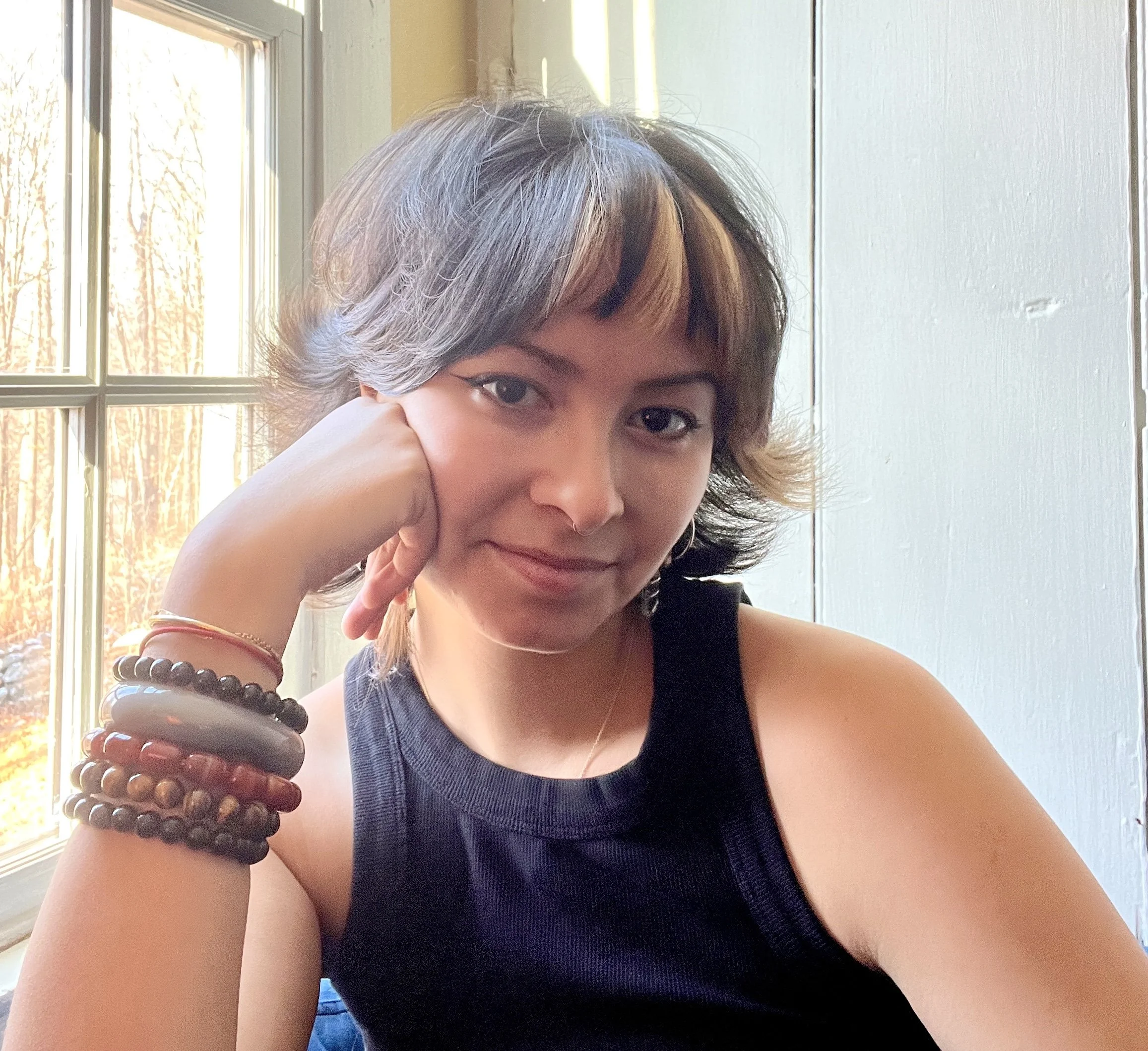 A woman with short, wavy hair that has blond highlights, wearing a black sleeveless top, resting her chin on her hand, and adorned with multiple colorful beaded bracelets on her wrist, sitting near a window with sunlight illuminating her face.
