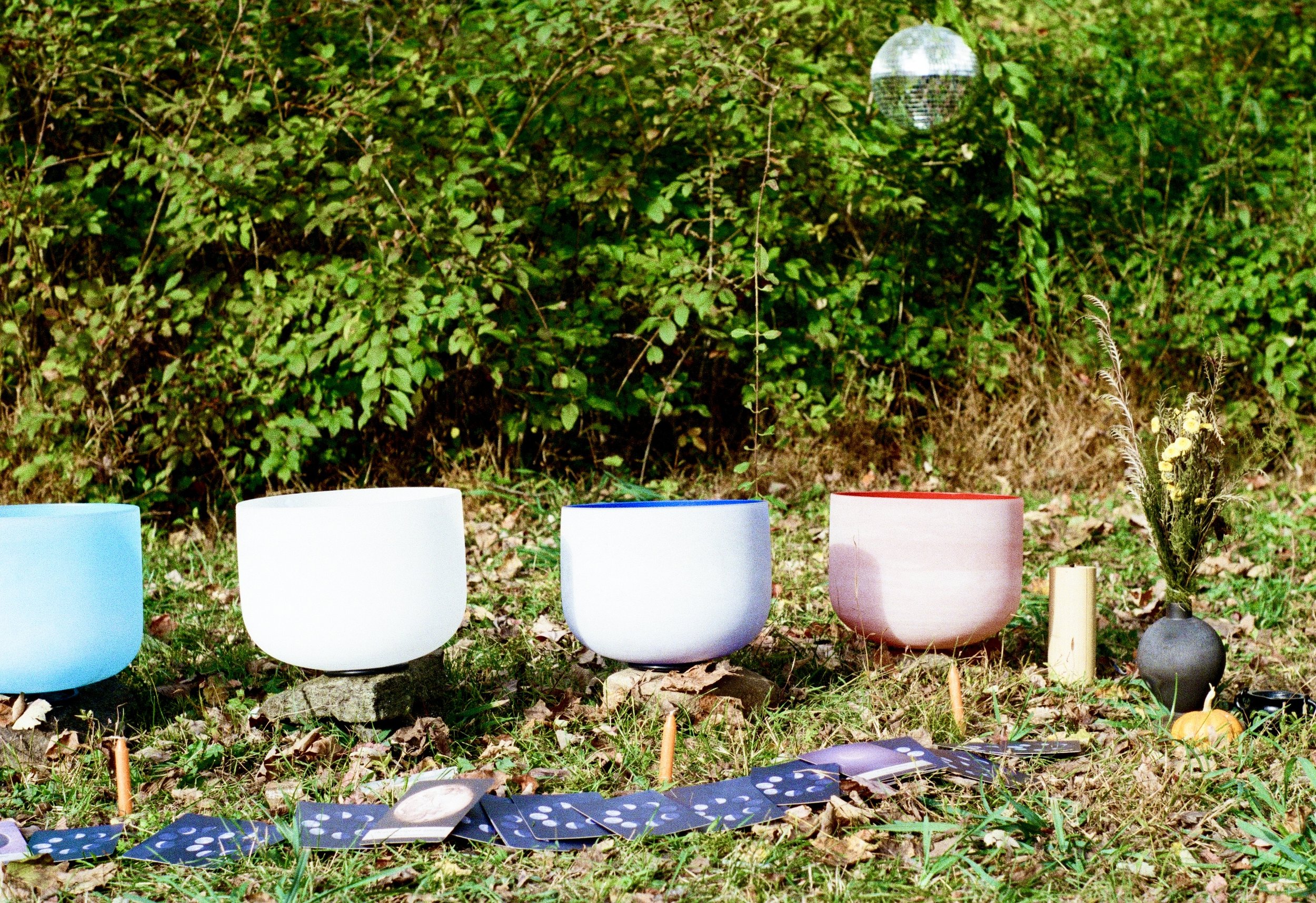 Colorful singing bowls arranged outdoors on grass with leaves, tarot cards, a small candle, a vase with dried flowers, and a small pumpkin, set against a background of green bushes and a small disco ball hanging in the trees.