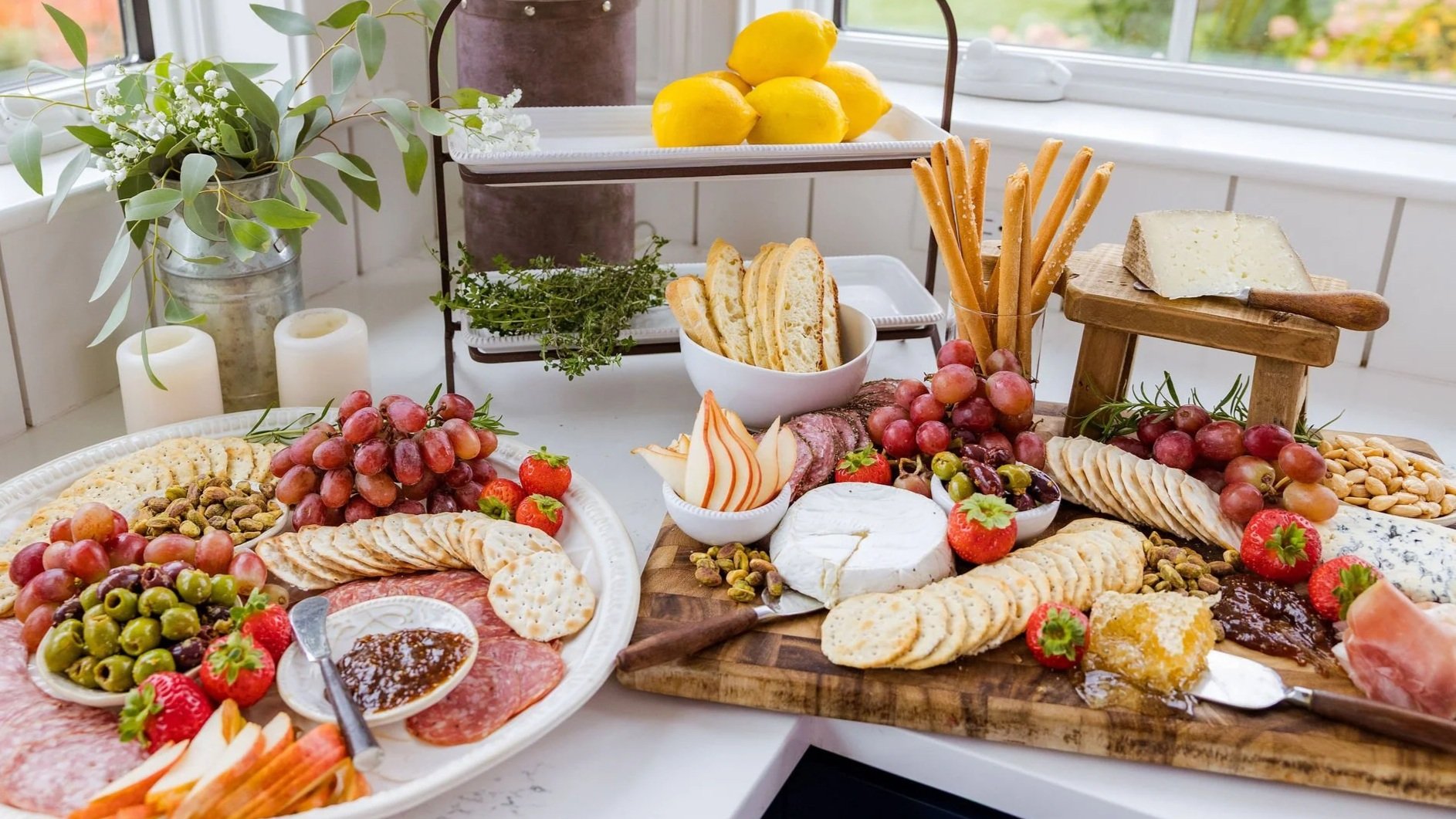 A spread of food including various cheeses, grapes, strawberries, olives, crackers, sliced meats, bread, and nuts on a white table, with a decorative tray of lemons and a plant in the background.