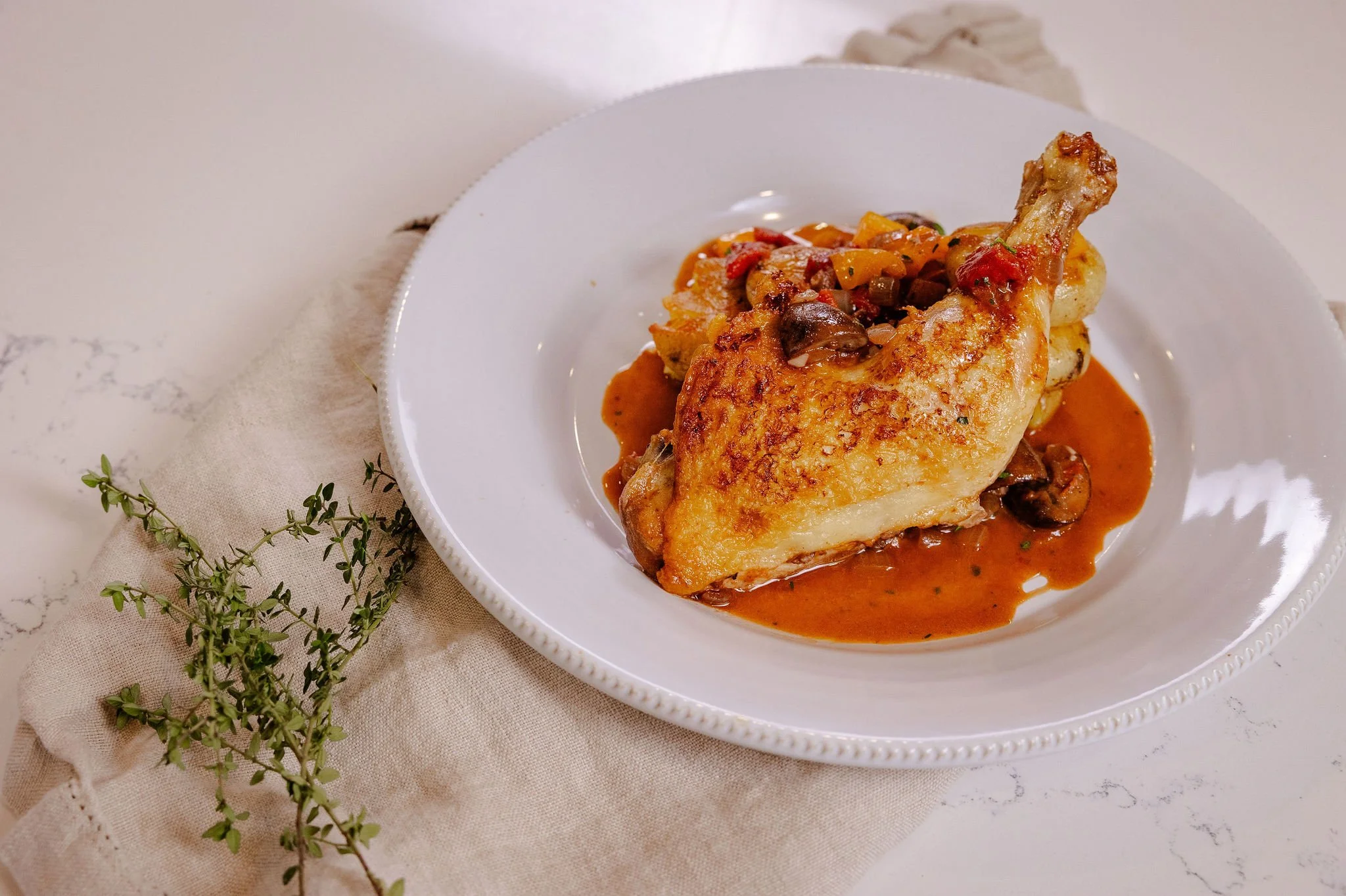 A roasted chicken leg in cacciatore sauce served on a white plate, sitting on a placement with fresh thyme.