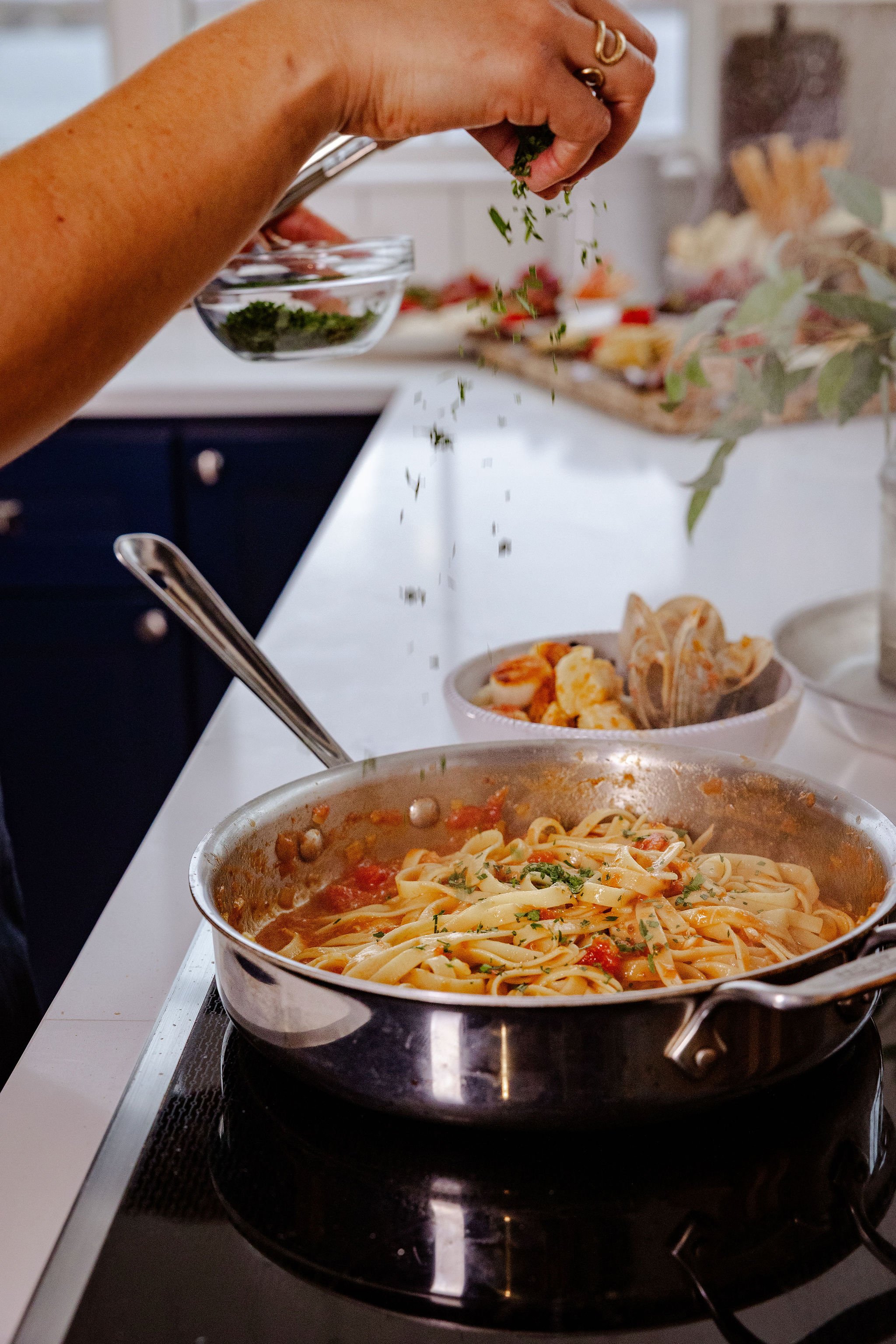 A person sprinkling chopped herbs onto a skillet of pasta with tomato sauce on a white stovetop. In the background, there are small bowls of additional ingredients and a bouquet of flowers.