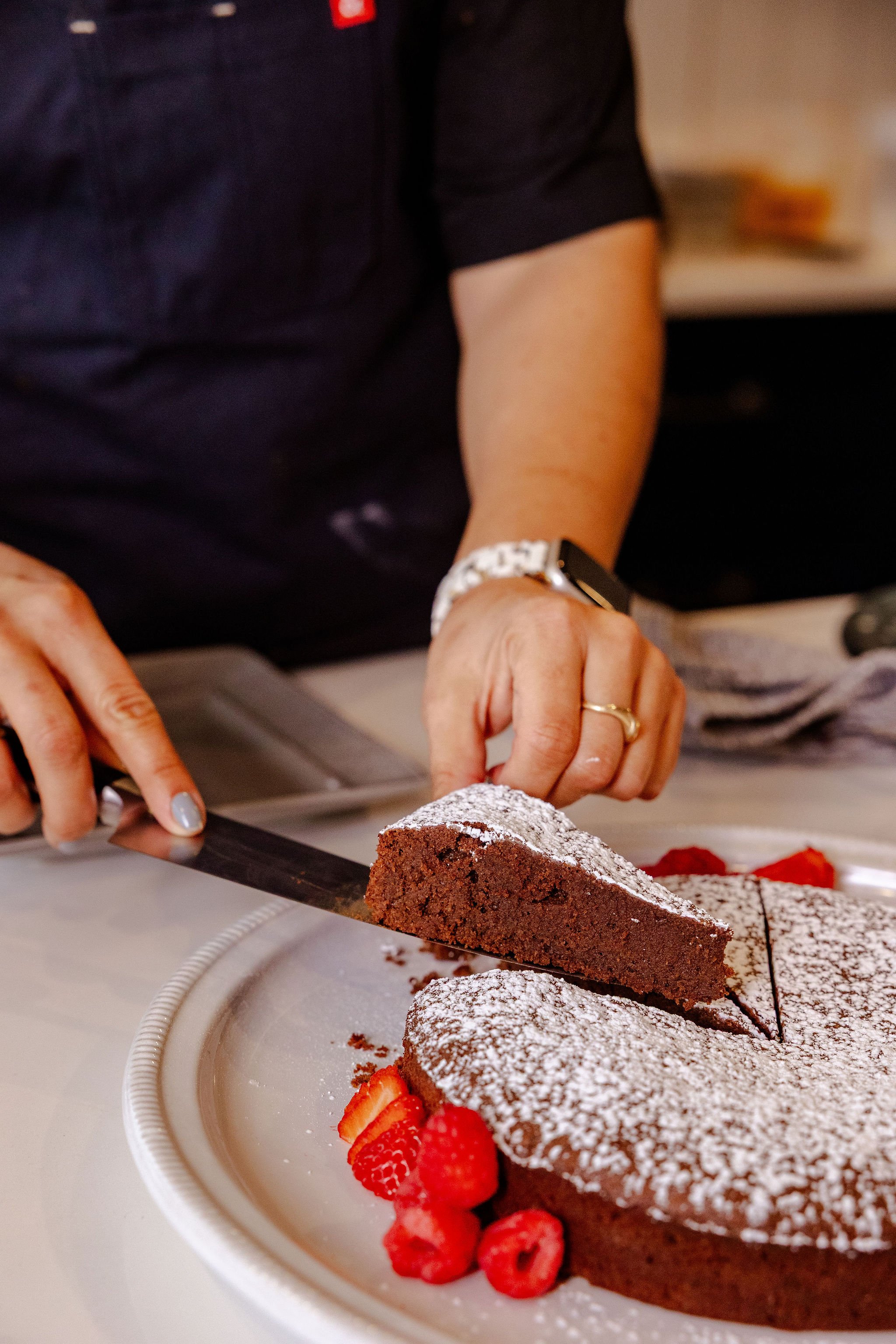 Chef Jenna Bethoney cutting a chocolate cake dusted with powdered sugar, garnished with strawberries on a white plate.