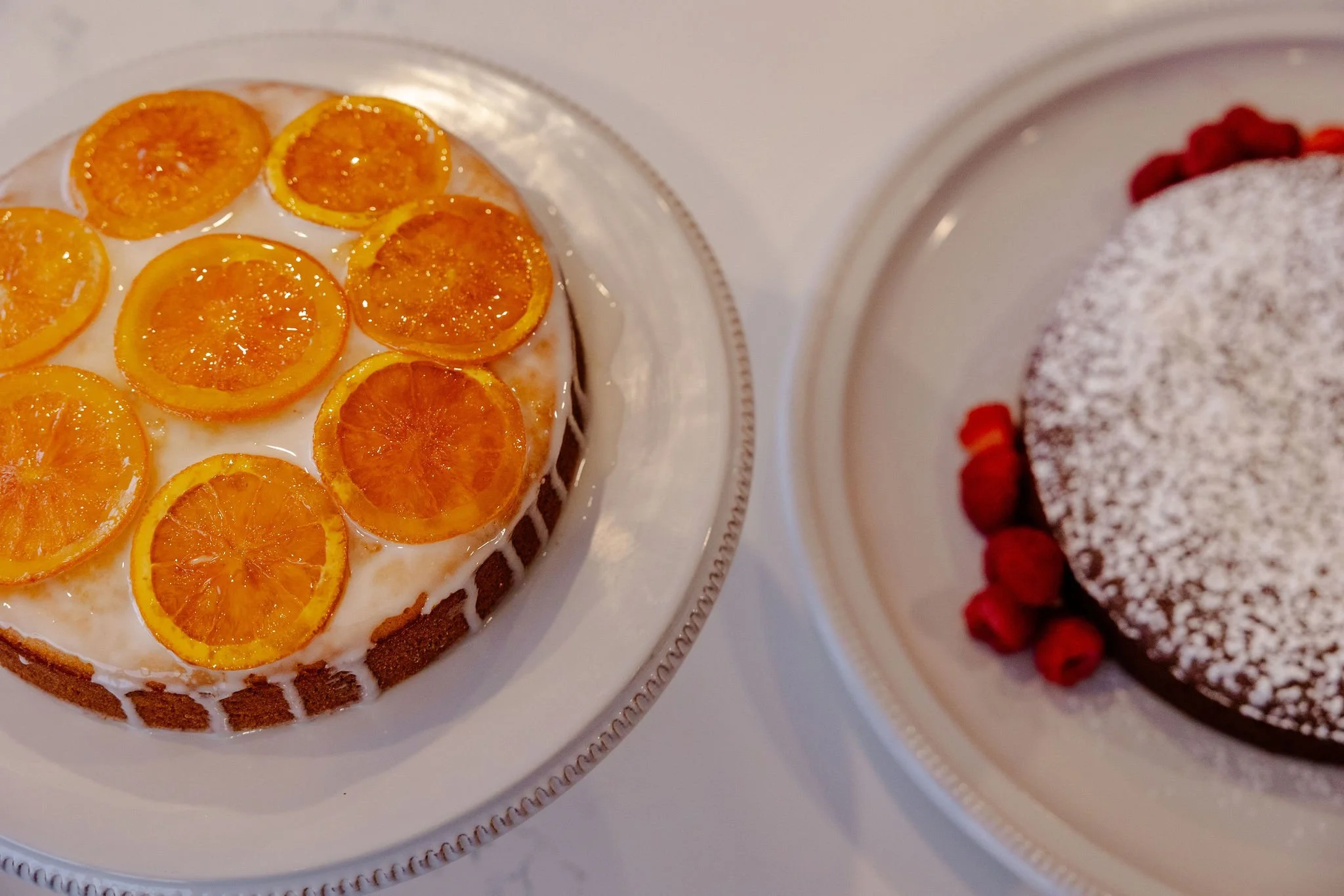 Two cakes on white plates; one is a citrus olive oil cake with orange slices on top, the other is a chocolate torte dusted with powdered sugar and garnished with red berries.