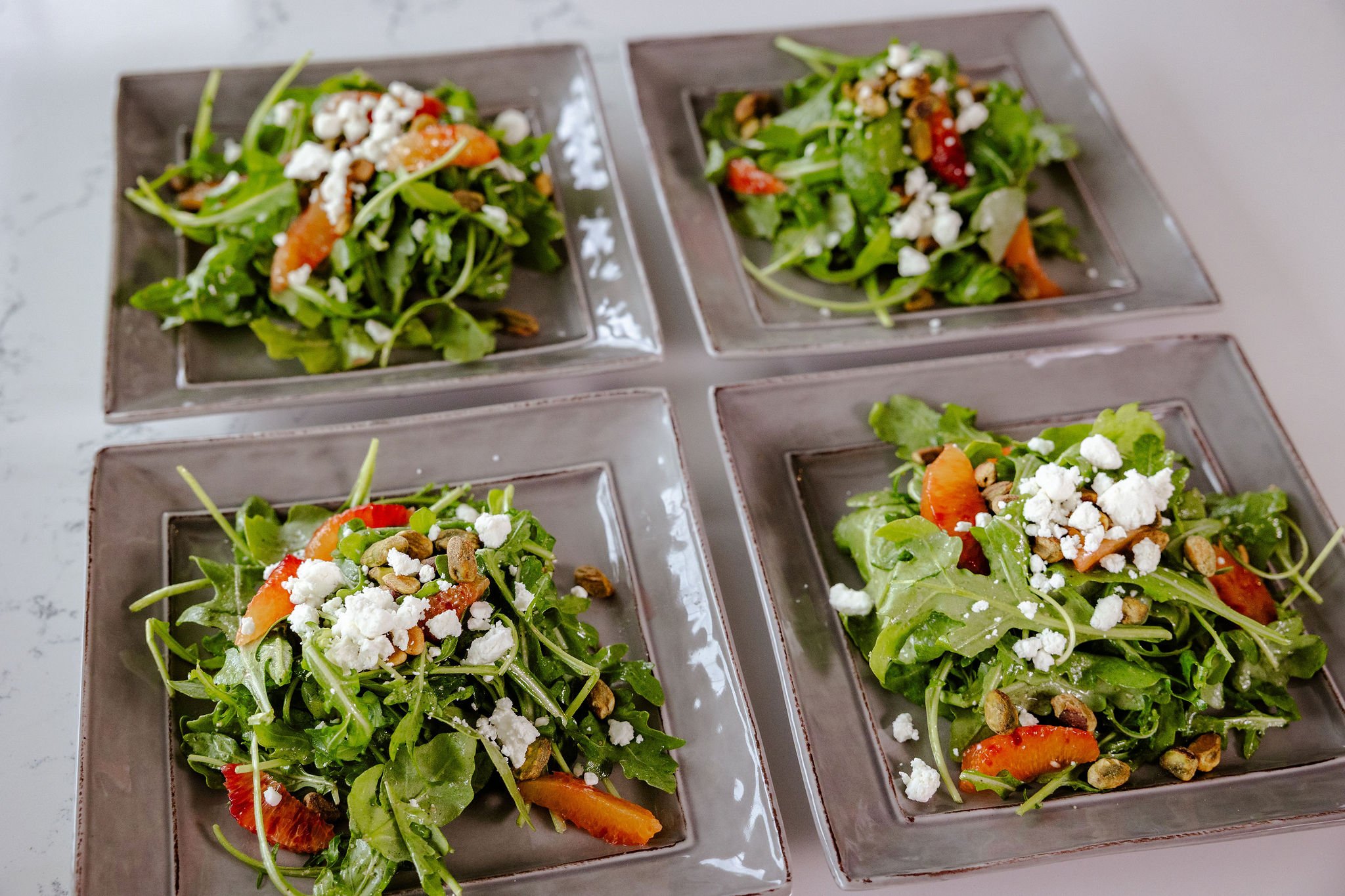 Four square plates of fresh green arugula salad topped with crumbled goat cheese, segmented blood orange, and toasted pistachios in a fennel seed vinaigrette, arranged on a beautiful surface.