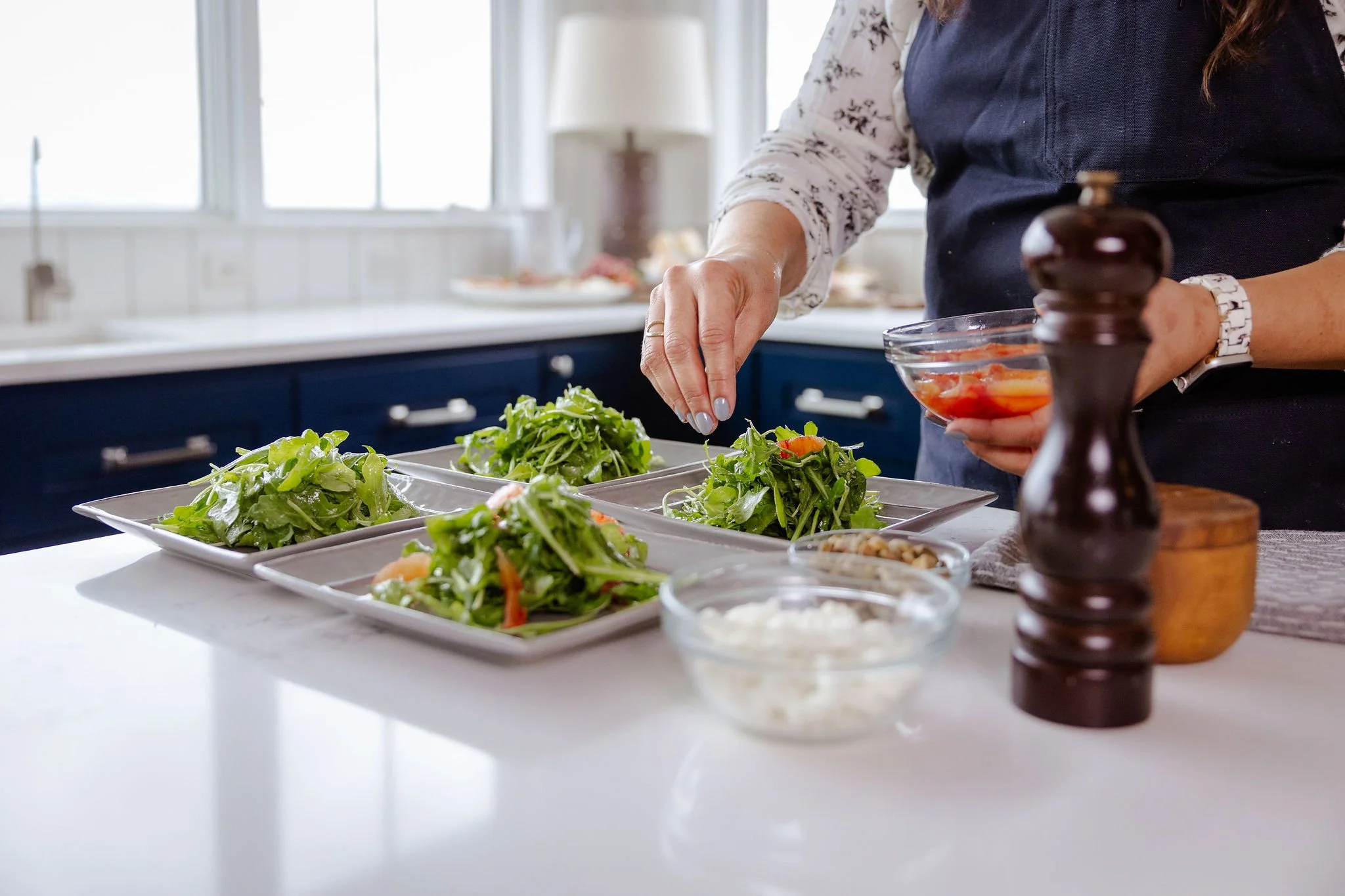 Chef Jenna Bethoney preparing baby arugula salad in a bright kitchen with blue cabinets, placing greens on square gray plates, with goat cheese and toasted fennel seed vinaigrette on the counter.