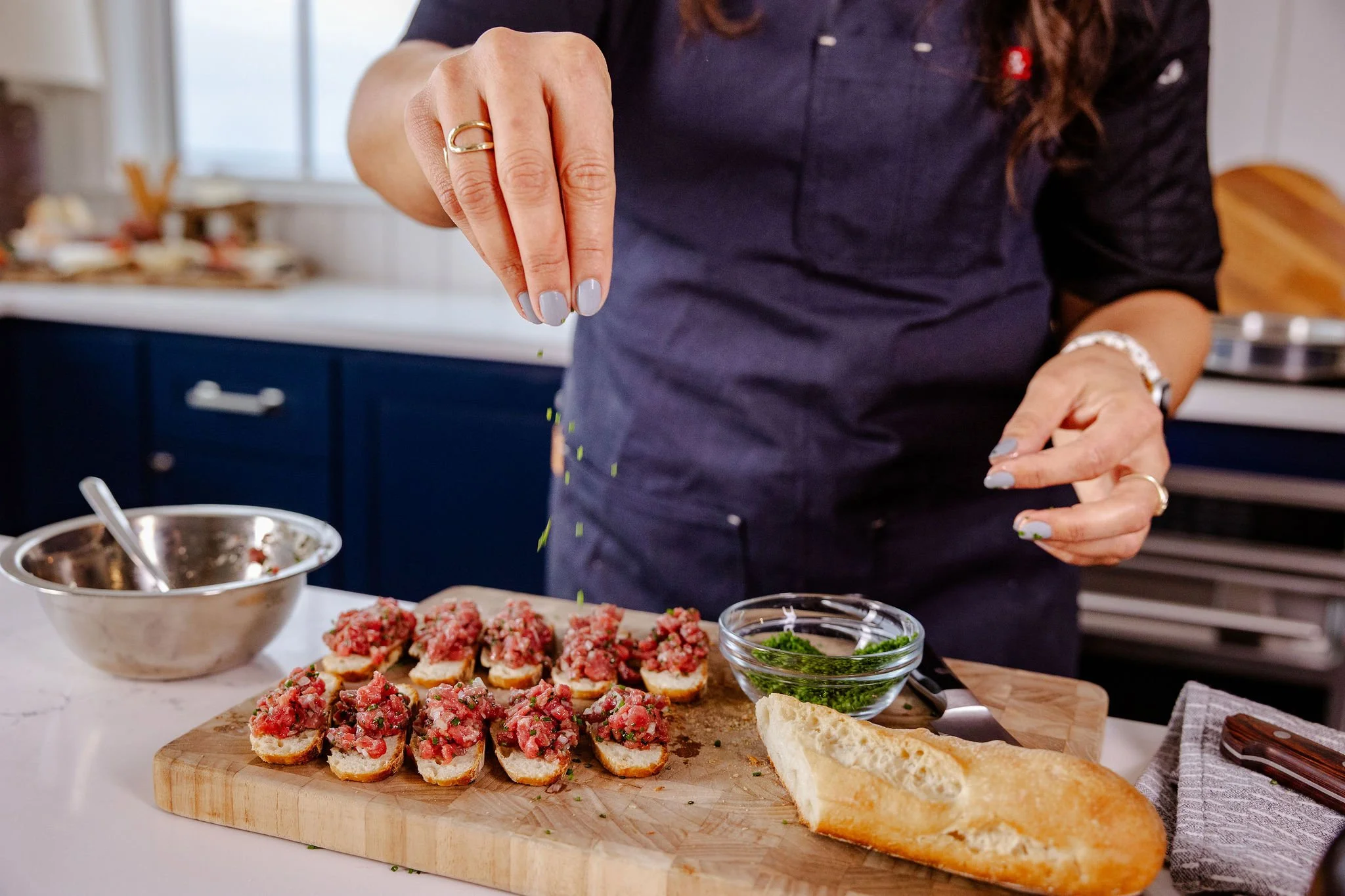 Chef Jenna Bethoney sprinkling chives on a tray of crostini topped with beef tartare, with bread and a small bowl of minced chives nearby in a kitchen.