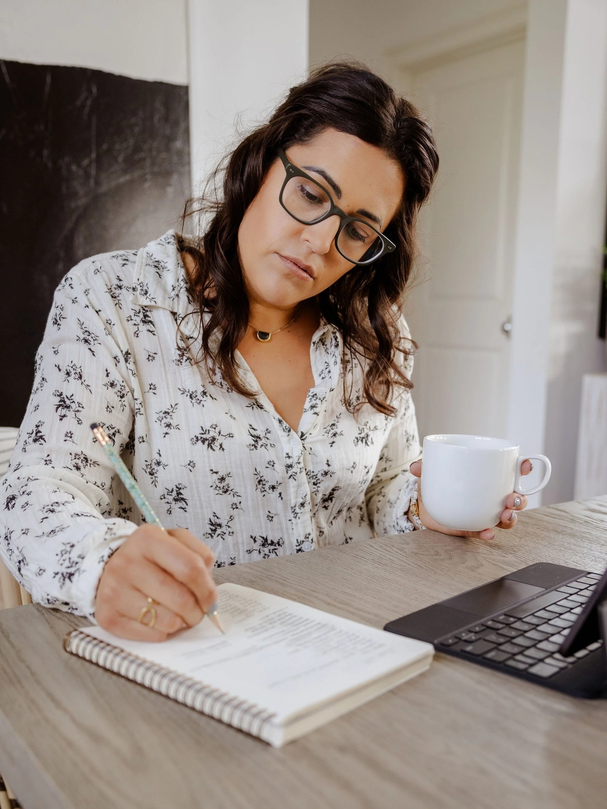 Chef Jenna Bethoney with dark hair, glasses, and wearing a white floral blouse, sitting at a wooden table, writing in a notebook, holding a coffee mug, with an iPad nearby in a bright room.