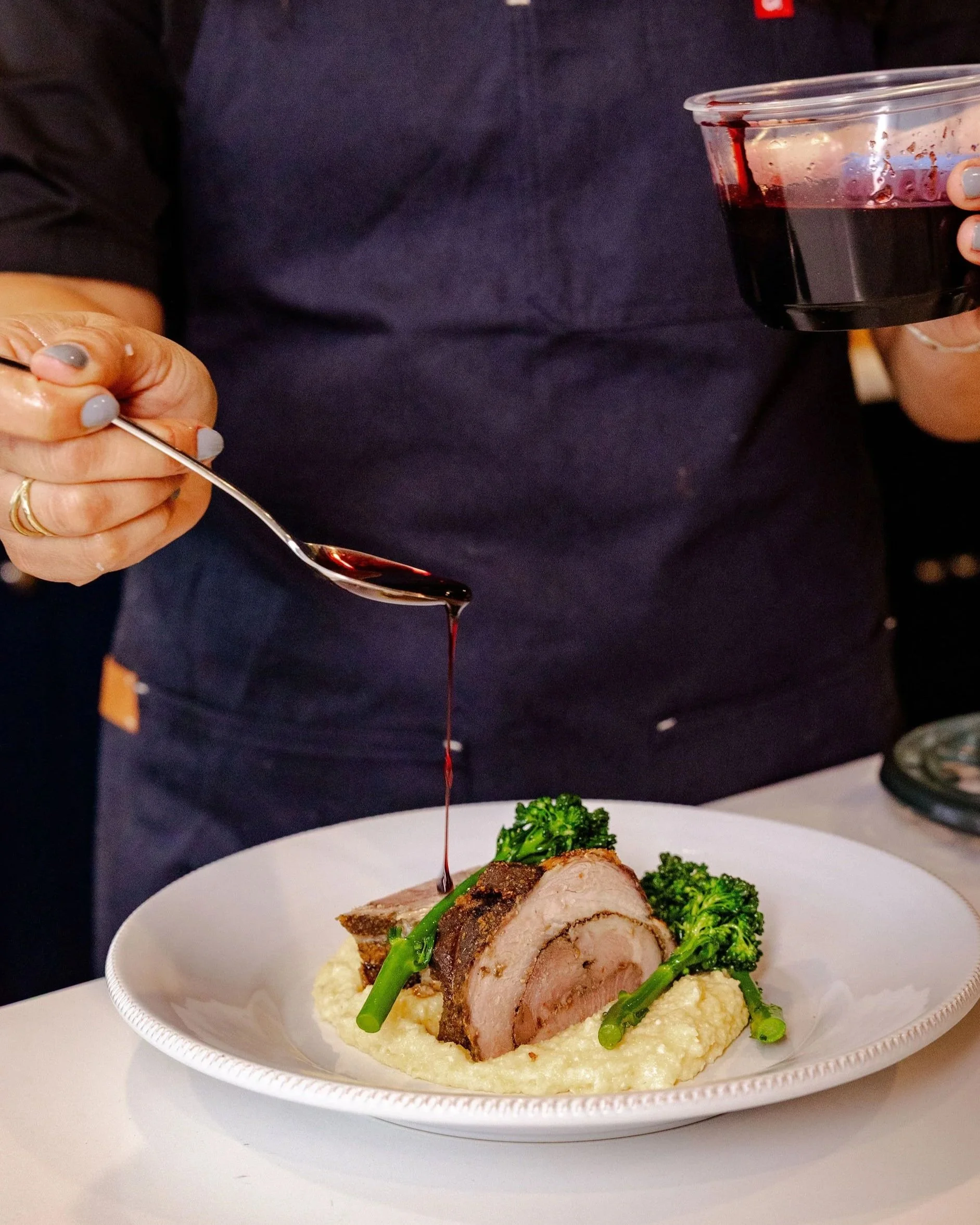 Person pouring red wine sauce over a plated dish of rolled pork tenderloin with broccoli and mashed potatoes.