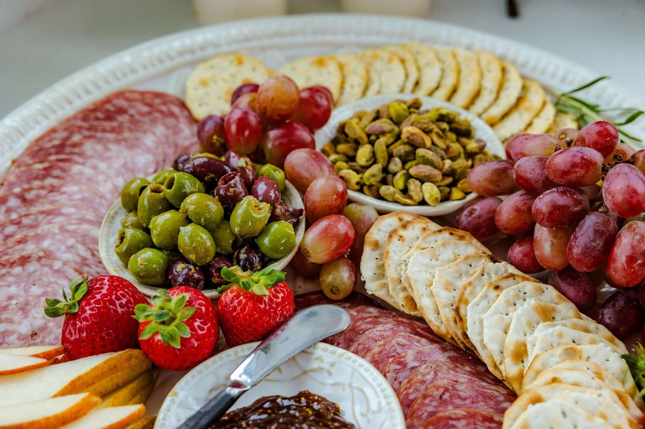 Charcuterie board with cured meats, marinated olives, cheeses, grapes, strawberries, fig preserves, pistachios, and crackers.