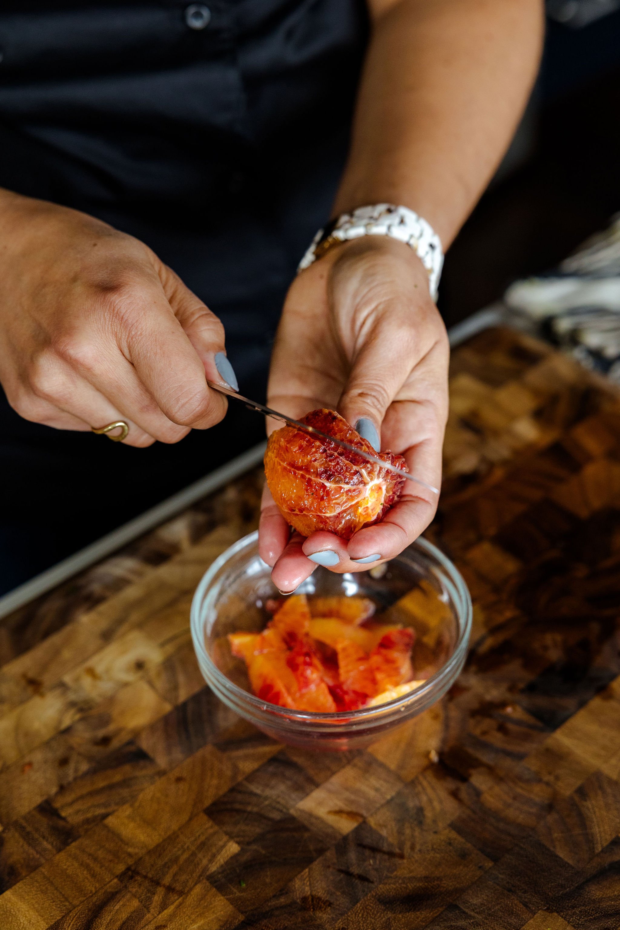 Chef Jenna Bethoney segmenting a blood orange over a glass bowl of blood orange segments on a wooden cutting board.