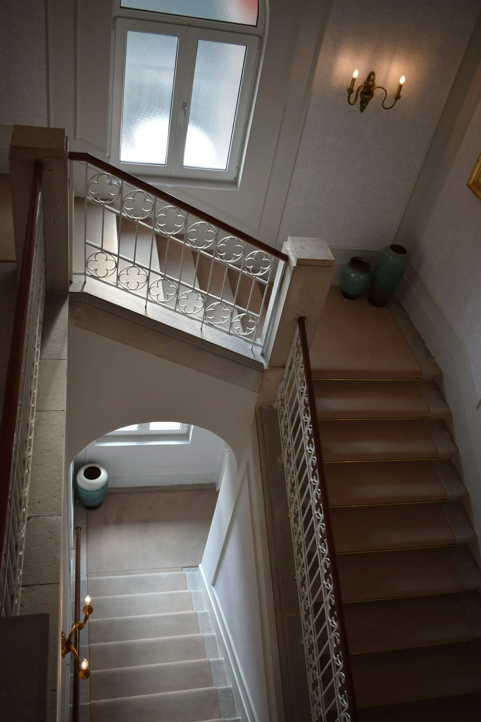Escalier intérieur avec balustrades en métal, vue du haut montrant un escalier descendant, deux vases verts dans un coin, une fenêtre au plafond, un Lustre avec deux bougies