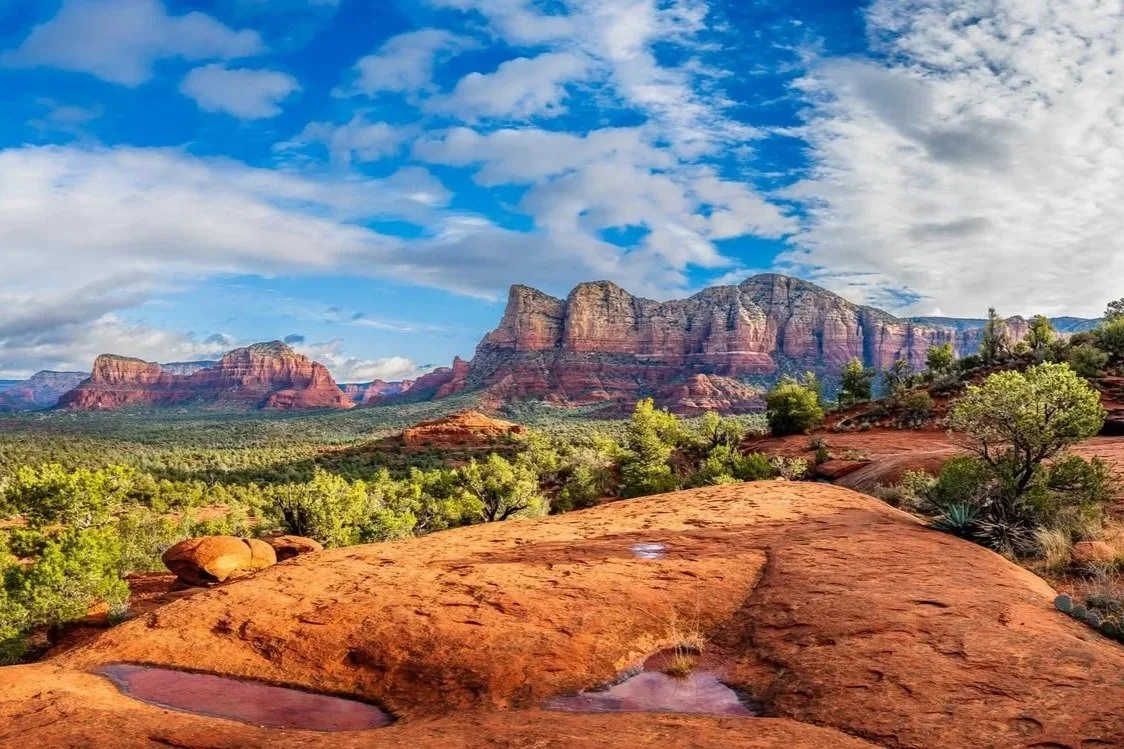 Wide-angle view of the iconic Sedona red rock formations under a blue sky with scattered clouds, captured from a premium slickrock overlook.