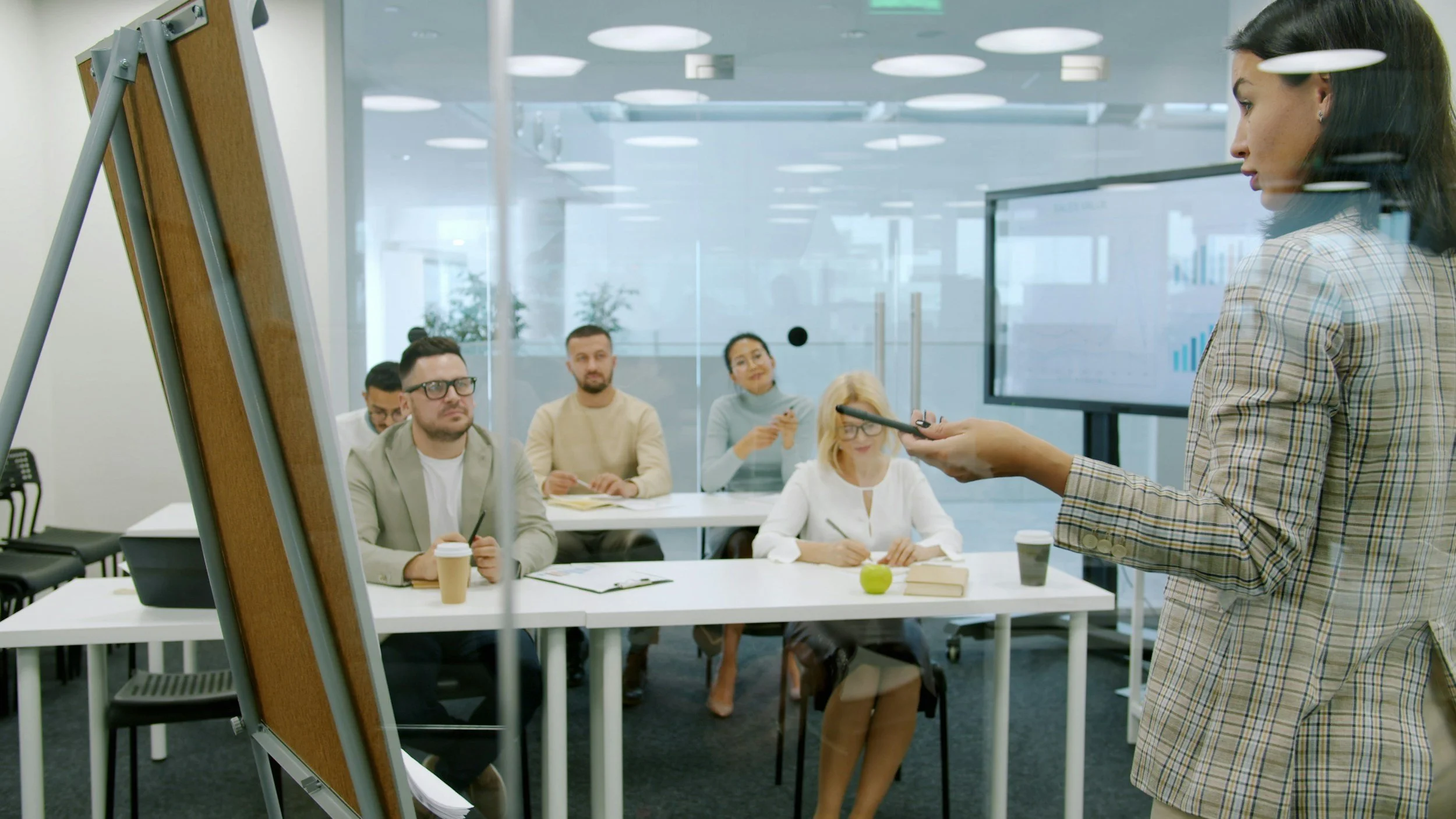 A woman giving a presentation to a group of five people seated at a conference table in a modern office, with a large screen displaying charts in the background.