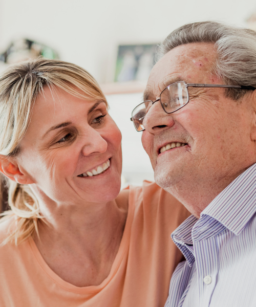 A smiling woman showing affection to an elderly man in glasses, both smiling happily in a close-up indoors.
