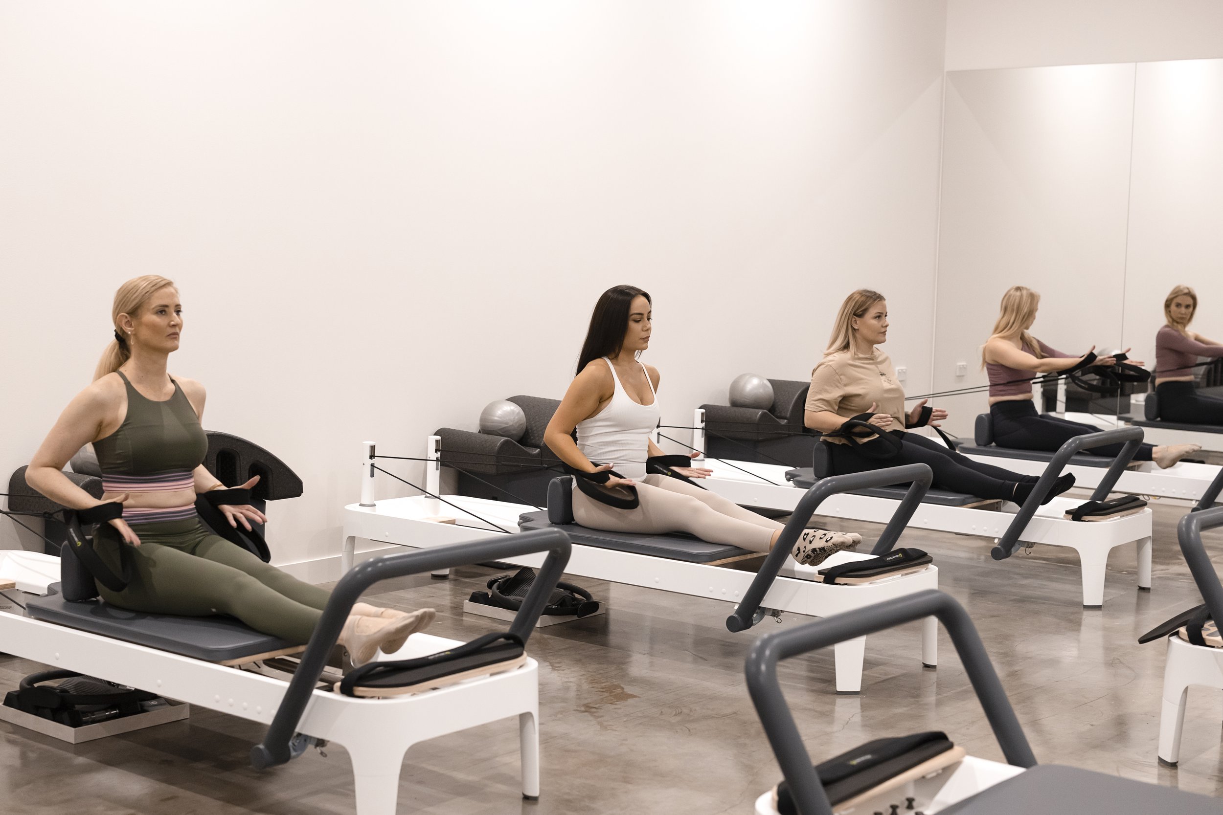 Four women seated on Pilates reformer machines in a fitness studio, engaging in a resistance workout with elastic bands around their wrists and ankles.