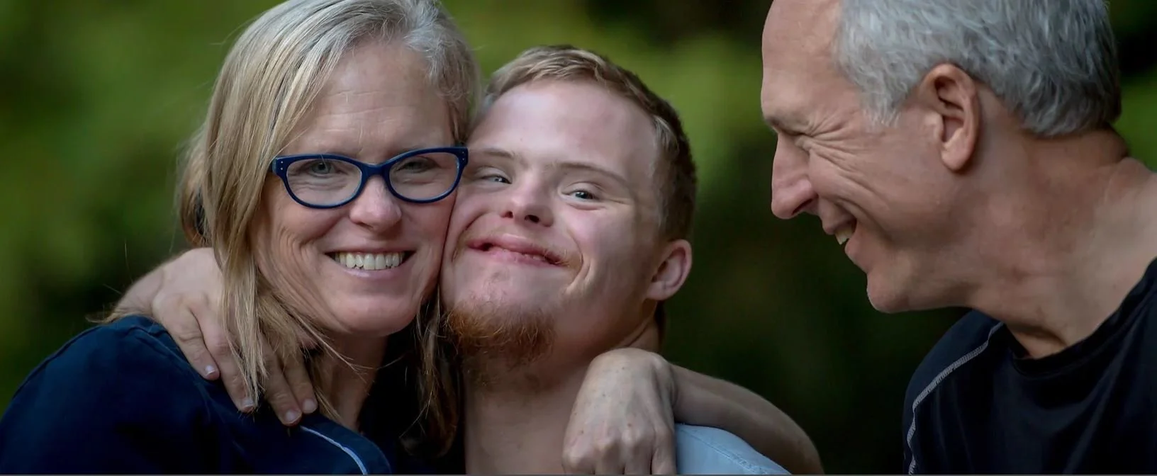 Three people smiling and hugging outdoors; a woman with glasses, a young man with short ginger hair and a beard, and an older man with gray hair.
