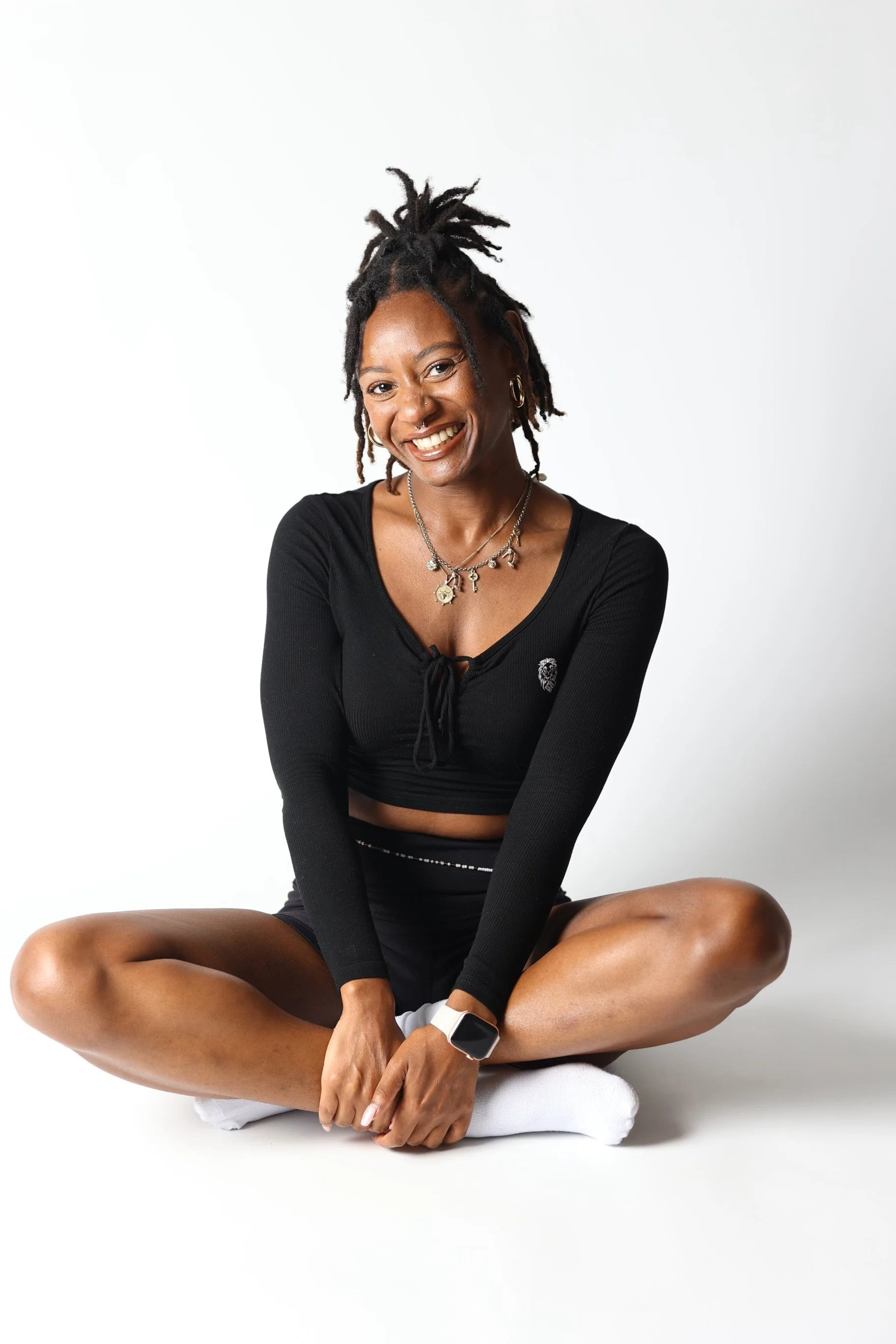 A woman with dreadlocks sitting cross-legged on the floor, smiling, wearing a black long-sleeve crop top, white socks, and a smartwatch.