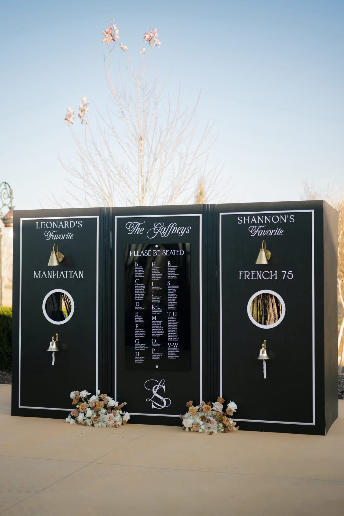 A black seating chart board for a wedding with vintage-style white lettering, floral decorations at the base, and brass wall lamps, displaying seating arrangements for the Gaffneys. Two signboards on either side show favorite items: Leonard's Manhattan and Shannon's French 75.