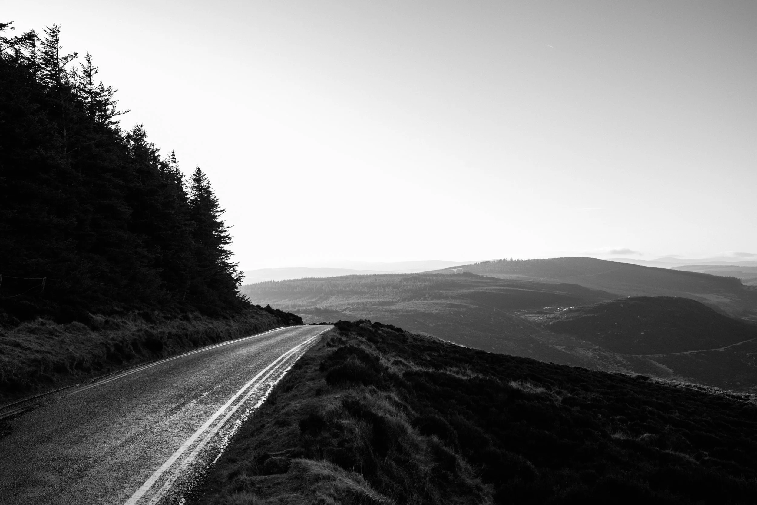 Een zwart-wit foto van een rechte weg die door een heuvelachtig landschap loopt, omringd door bomen en open velden onder een heldere hemel.