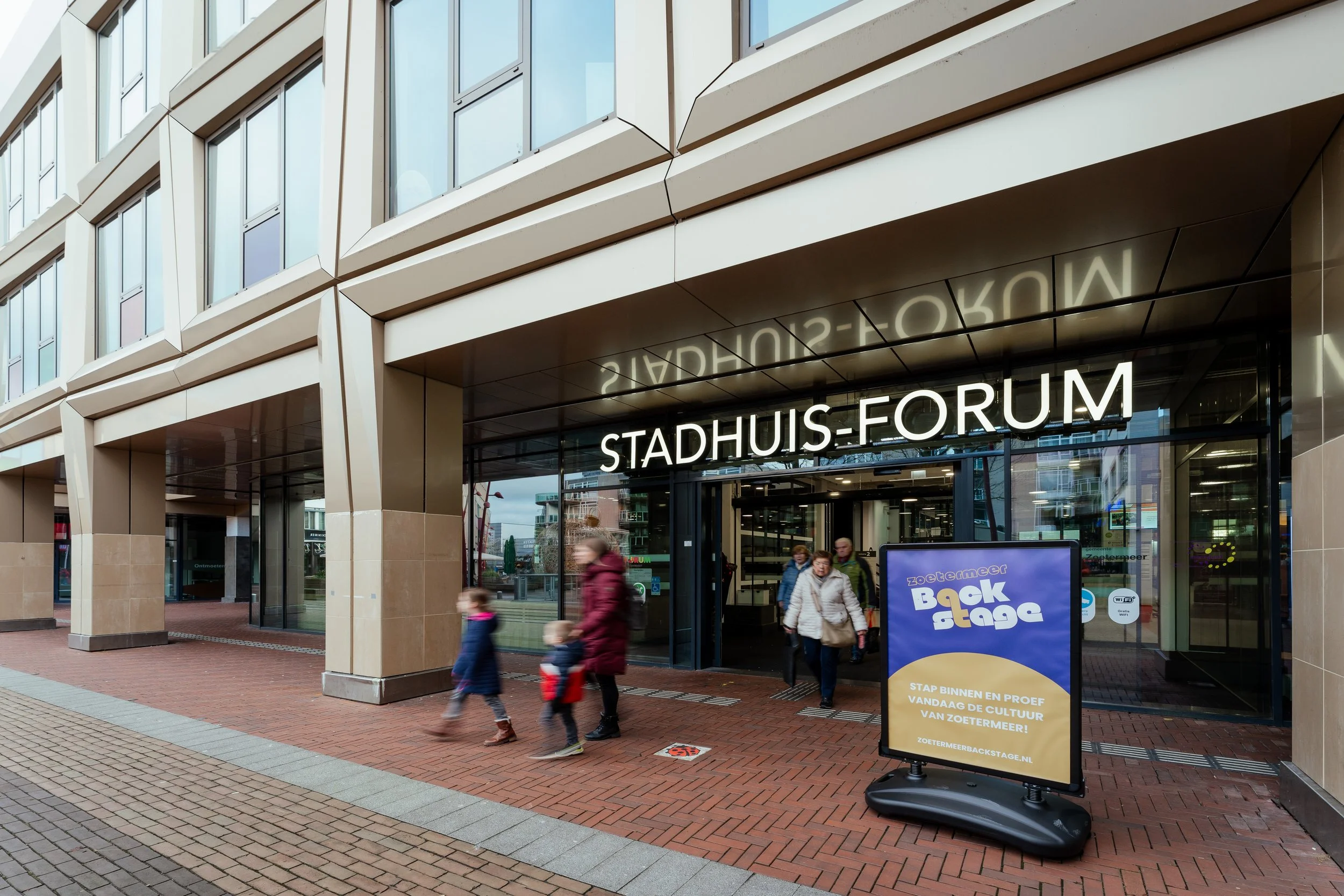 De ingang van het Stadshuis-Forum met mensen die naar binnen en buiten lopen, een pijlwagen met een reclamebord ernaast en een modern gebouw met grote ramen.