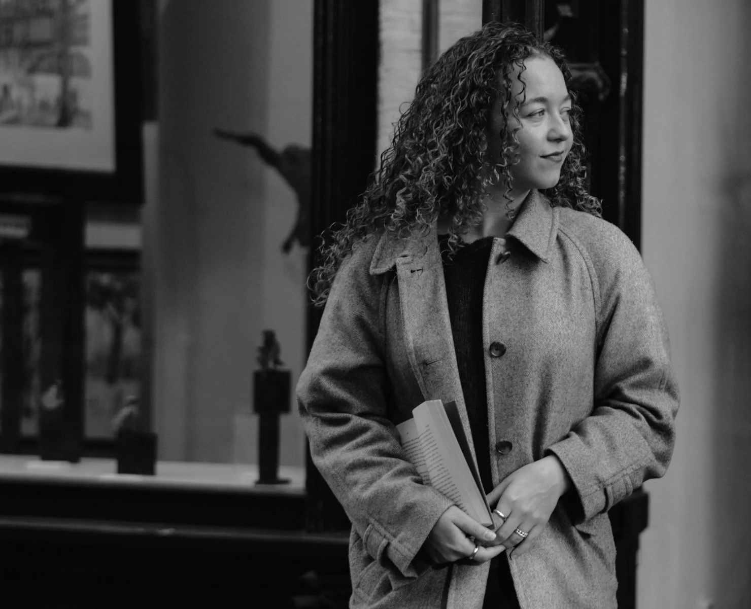 Een vrouw met krullend haar draagt een jas en houdt boeken vast, zij kijkt naar rechts in een kunstgalerij of museum met beeldhouwwerken op de achtergrond, zwart-wit foto.